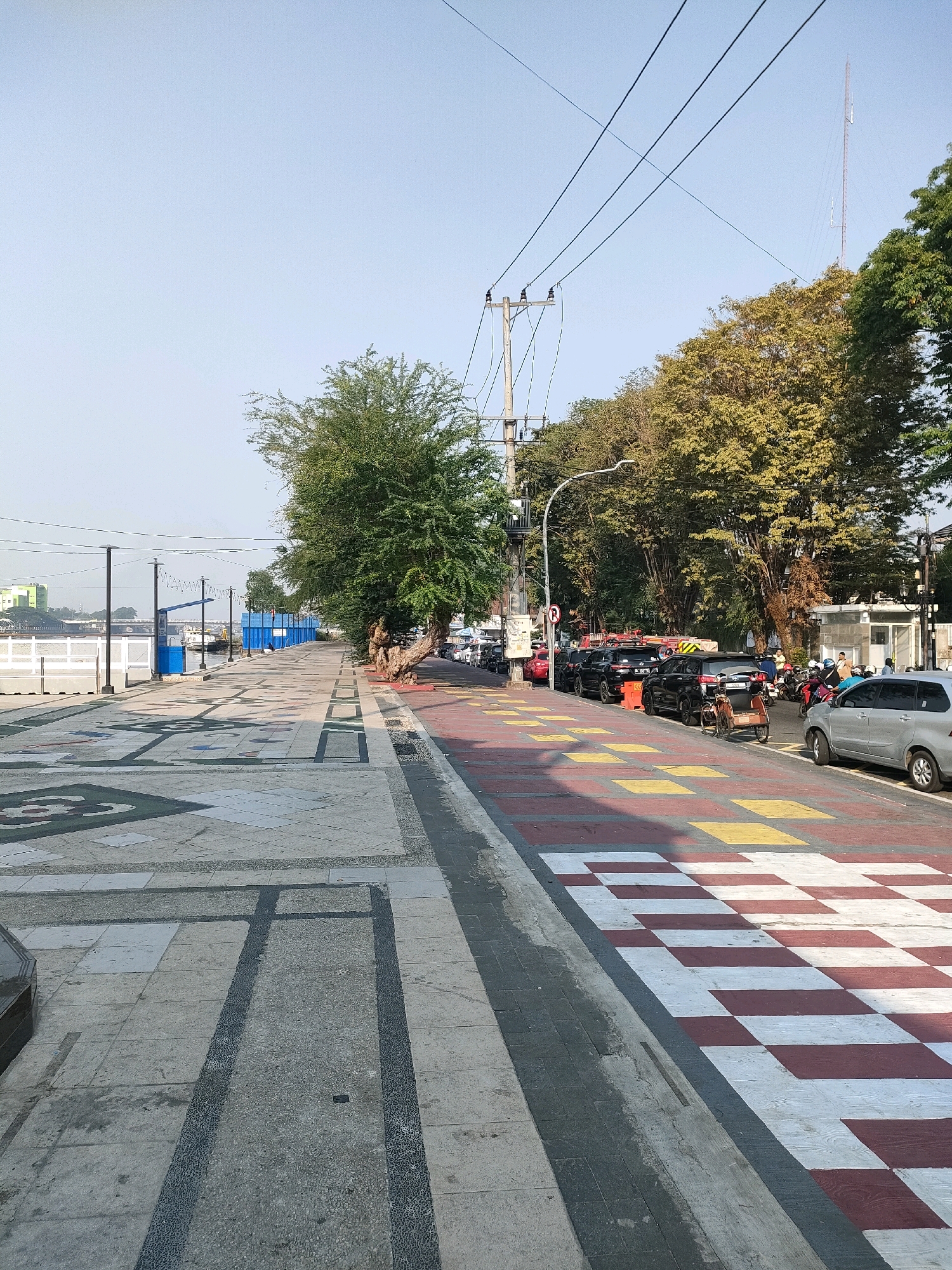 The image depicts a pedestrian walkway alongside a street in Banjarmasin, Indonesia. The walkway is paved with patterned tiles, featuring geometric designs. To the left of the walkway, a body of water is visible, with a white barrier along its edge and some buildings in the distance. A line of trees with green and yellow foliage runs along the right side of the walkway, bordering a street. The street is lined with parked cars, including several black SUVs and a silver minivan. A few motorcycles and a three-wheeled vehicle are also present. Power lines and a tall radio tower are visible in the sky. The scene appears to be in daylight, with clear skies.