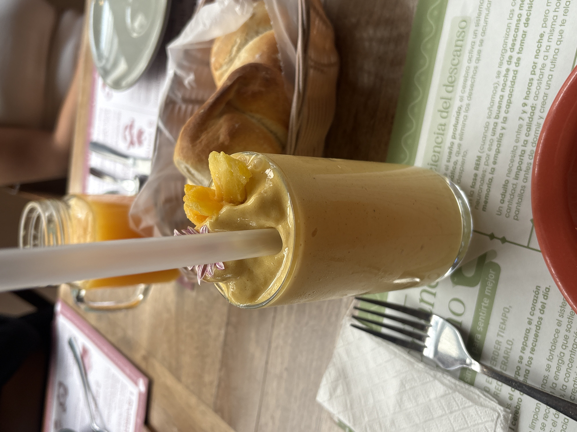 A light brown smoothie with a yellow fruit garnish and a white straw sits horizontally on a wooden table next to a fork. The table is also set with a basket of bread, a small glass jar of orange juice, and a plate. A newspaper placemat is visible under the smoothie and fork.