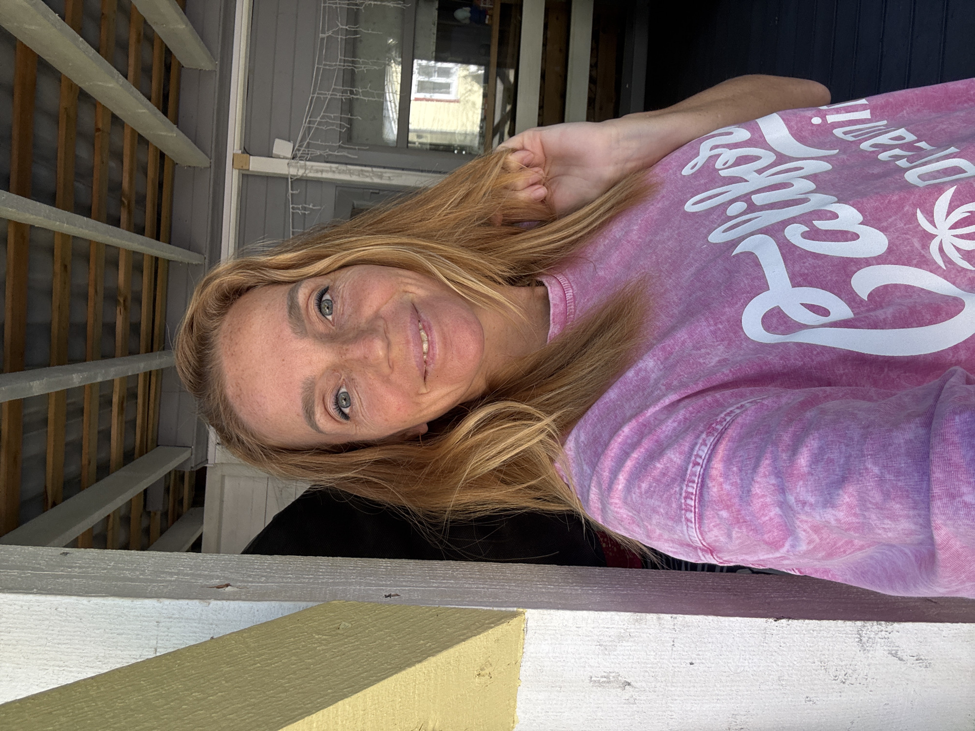 A woman with light brown hair and freckles smiles at the camera, holding a strand of her hair. She is wearing a pink tie-dye t-shirt with the words "California Dreaming" and a palm tree graphic printed in white. The woman is standing in an outdoor, covered area, possibly a porch or deck, with a wooden ceiling visible above. To her left, a white wooden beam and a yellow-painted beam are prominent. To her right, a grey siding wall and a glass-paned window are visible, reflecting a yellow building. The floor appears to be dark wood planks. The location is not identifiable as Zelenaya Roshcha, Russia.