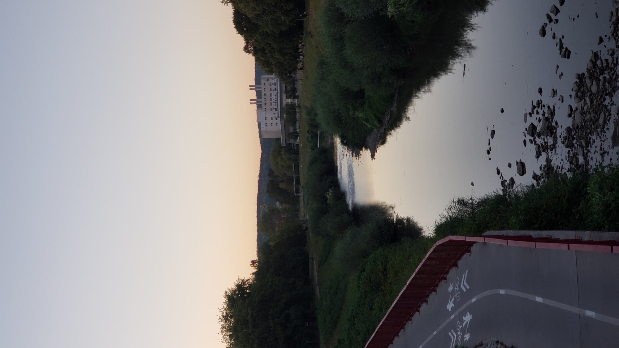 The image shows a river flowing through a park-like area in the city of Prešov, Slovakia. On the left side of the river, there is a paved pathway with a red railing, marked with symbols for bicycles and pedestrians. Lush green trees and bushes line the banks of the river. In the background, across the river, a large, modern building with multiple smokestacks is visible, set against a backdrop of rolling hills. The sky above is a soft gradient of pale blue and orange, suggesting either sunrise or sunset. The water of the river is calm and reflects the sky and surrounding greenery. Rocks are scattered along the riverbed and its edges. Several people are faintly visible in the distance on the right bank, near the building, suggesting a recreational area.