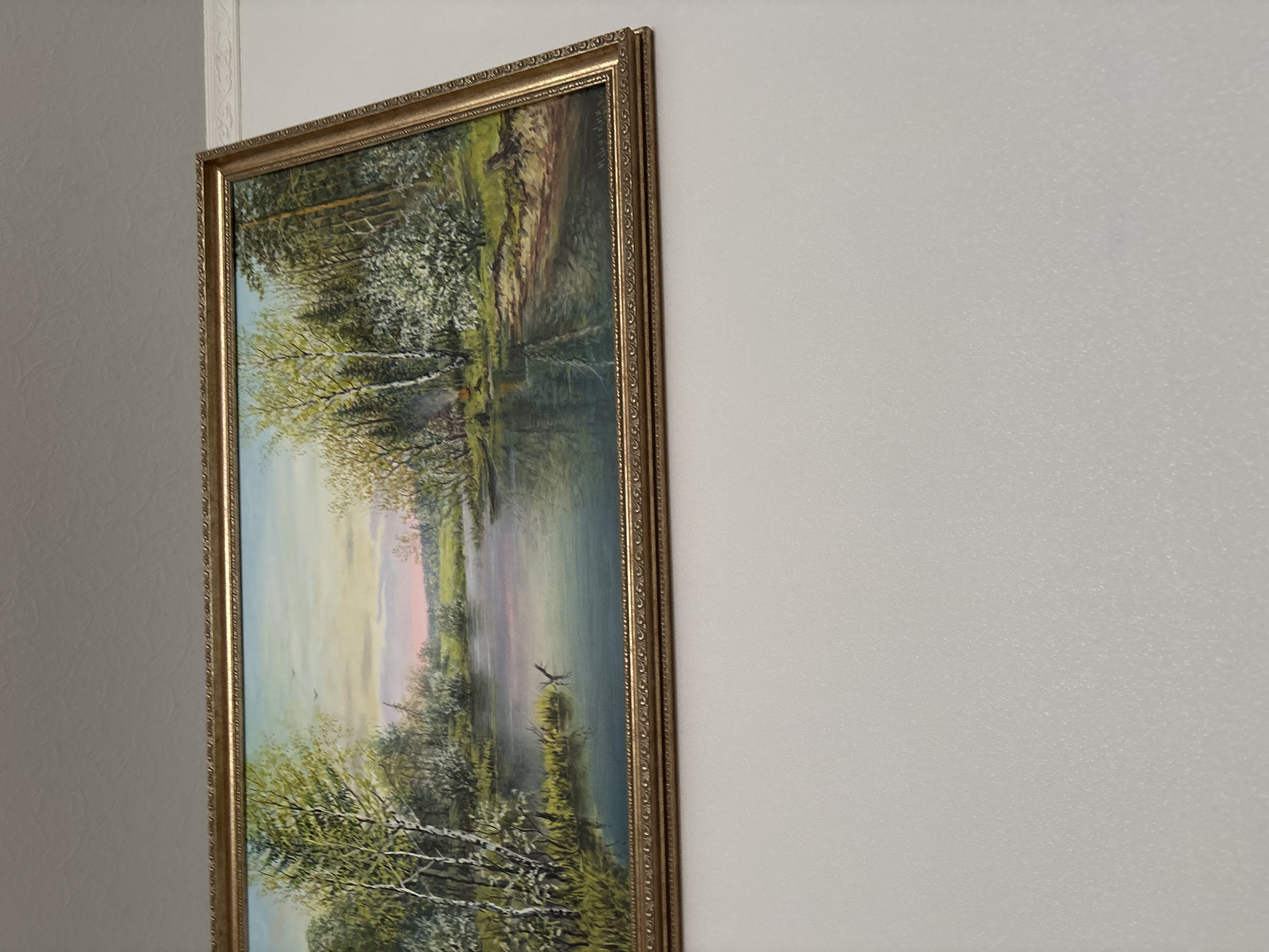 A framed painting of a landscape hangs on a white wall. The painting depicts a tranquil scene with trees reflected in a body of water, likely a lake or river. The sky is visible with hues of blue, yellow, and pink. The frame is ornate and gold in color. The environment is likely an interior space, based on the wall. There is no information to determine if this image relates to Ferma, Russia.
