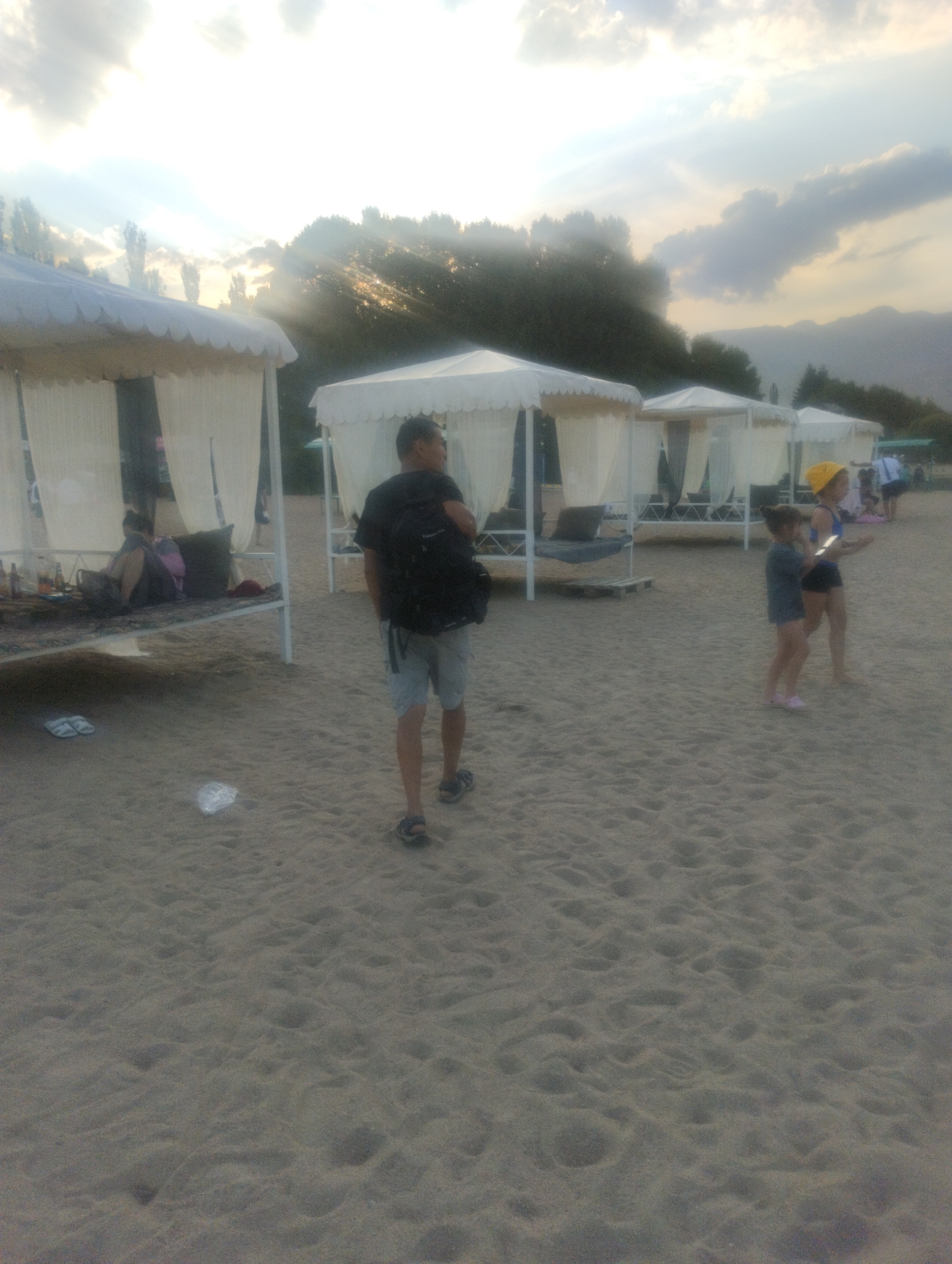 The image depicts a beach scene in Cholpon-Ata, Kyrgyzstan, characterized by sand and a cloudy sky with sun rays piercing through. Several white cabanas with curtains are set up on the sand, offering shaded resting areas. In the foreground, a man wearing a black t-shirt, grey shorts, and sandals is walking away from the camera, carrying a backpack. Further in the background, two young girls are visible on the sand, one wearing a yellow hat and a blue top, the other in a grey top and dark shorts. A person is seated in one of the cabanas on the left, partially obscured. The beach appears to be part of a resort or recreational area, with trees and mountains forming the backdrop. The overall impression is a casual, outdoor setting, likely during daylight hours.