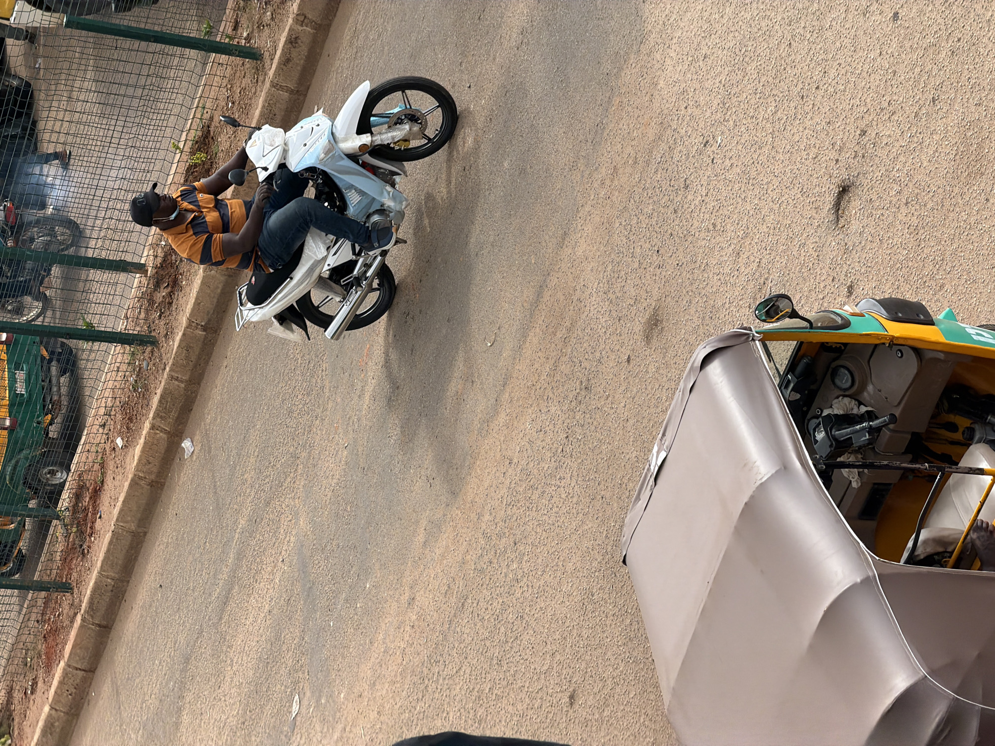 In Bukuru, Nigeria, a person wearing a striped shirt and dark pants rides a white motorcycle. The motorcycle has a light blue plastic covering on its front fairing. The person is wearing a black baseball cap and a face mask pulled down below their chin. They are riding on a paved road with a curb on the left side. A portion of a beige-colored auto-rickshaw is visible in the foreground, its roof is slightly dented. In the background, a metal fence separates the road from an area with some vegetation and what appears to be a green vehicle. The lighting suggests it is daytime, with shadows cast on the road.