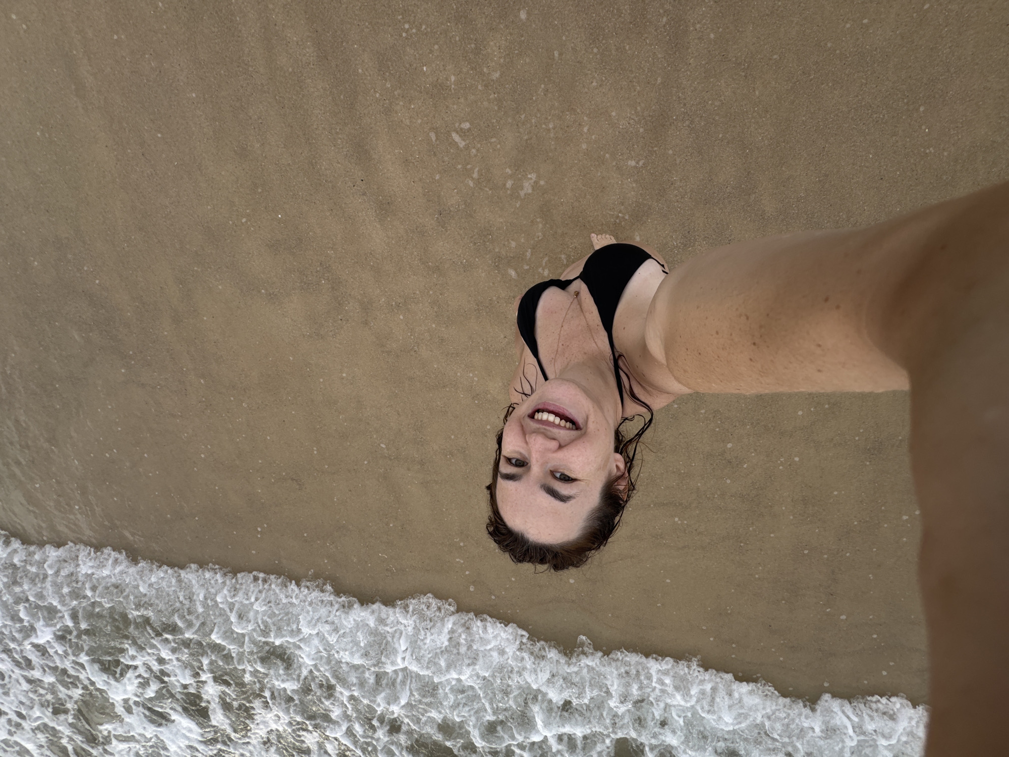 A woman is taking a selfie at the beach. She is wearing a black bikini and a necklace. She is smiling at the camera. Her hair is wet and appears to be dark brown. The beach has light brown sand and the ocean waves are breaking on the shore. The water is clear and foamy. The lighting suggests it is daytime, likely with overcast skies or late afternoon sun. The context provided indicates the location is Choeng Thale, Thailand. The woman's expression is joyful and relaxed, indicating she is enjoying her time at the beach.