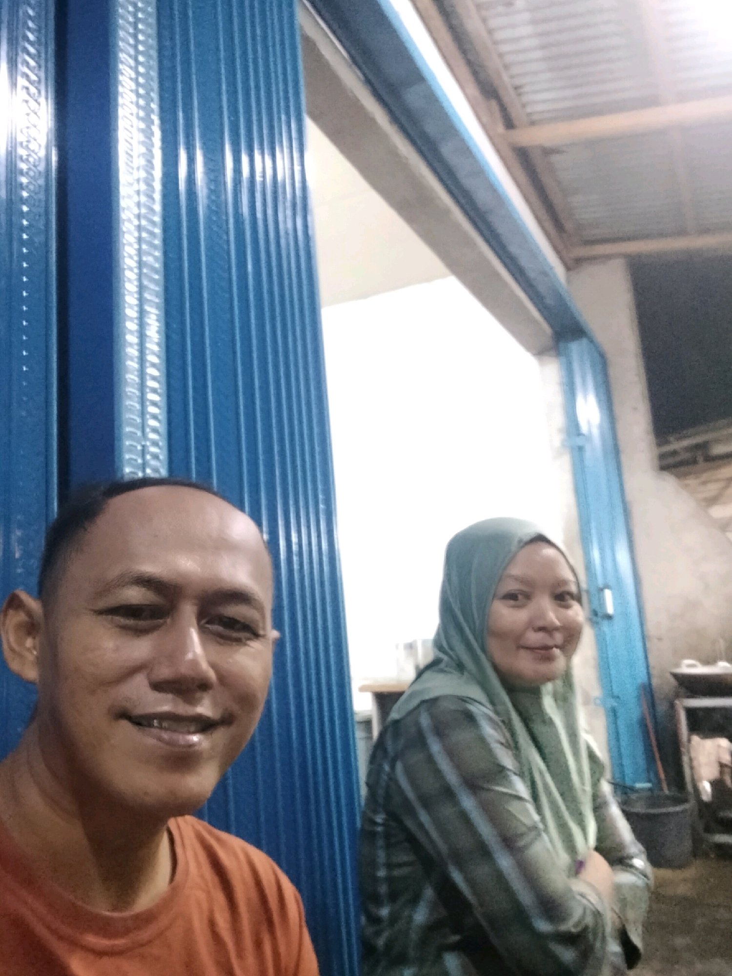 In the city of Arang Limbung, Indonesia, a close-up selfie shows a man with short hair and an orange t-shirt in the foreground, smiling at the camera. To his right, a woman wearing a light green hijab and a striped garment is also visible, smiling. They are positioned in front of a blue corrugated metal door, which appears to be an entrance to a building. The background is dimly lit and suggests an outdoor or semi-enclosed space, possibly a shop or a dwelling, with hints of corrugated metal roofing and various objects in the periphery.