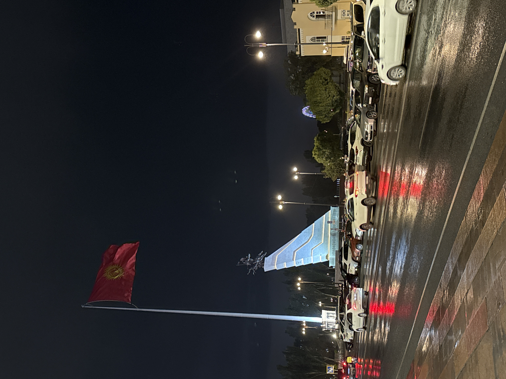 The image captures Ala-Too Square in Bishkek, Kyrgyzstan, at night. A large flag of Kyrgyzstan flies prominently on a tall flagpole on the left side of the frame. To the right of the flagpole, a prominent monument features a statue of horsemen atop a stepped, illuminated structure. The street in the foreground is wet, reflecting the red and white lights of various vehicles, suggesting recent rain or wet conditions. A line of parked cars and vehicles in motion are visible on the street, with streetlights illuminating the scene. In the background, trees are visible, and further back, a Ferris wheel with colorful lights can be seen, along with a light-colored building. The overall atmosphere is urban and nocturnal.