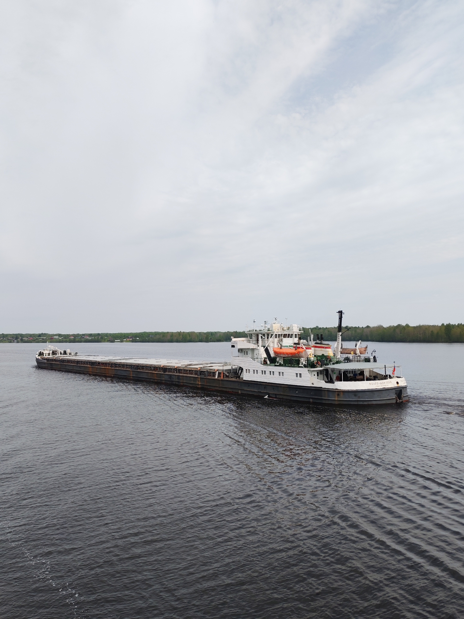 A barge is moving on the water in the area of Раменье, Russia. The barge is gray and white, with a long flat deck and a white superstructure. It is sailing on a river or lake under an overcast sky. The shoreline is visible in the distance, with trees and some buildings. The water is dark and rippled, indicating a light wind.