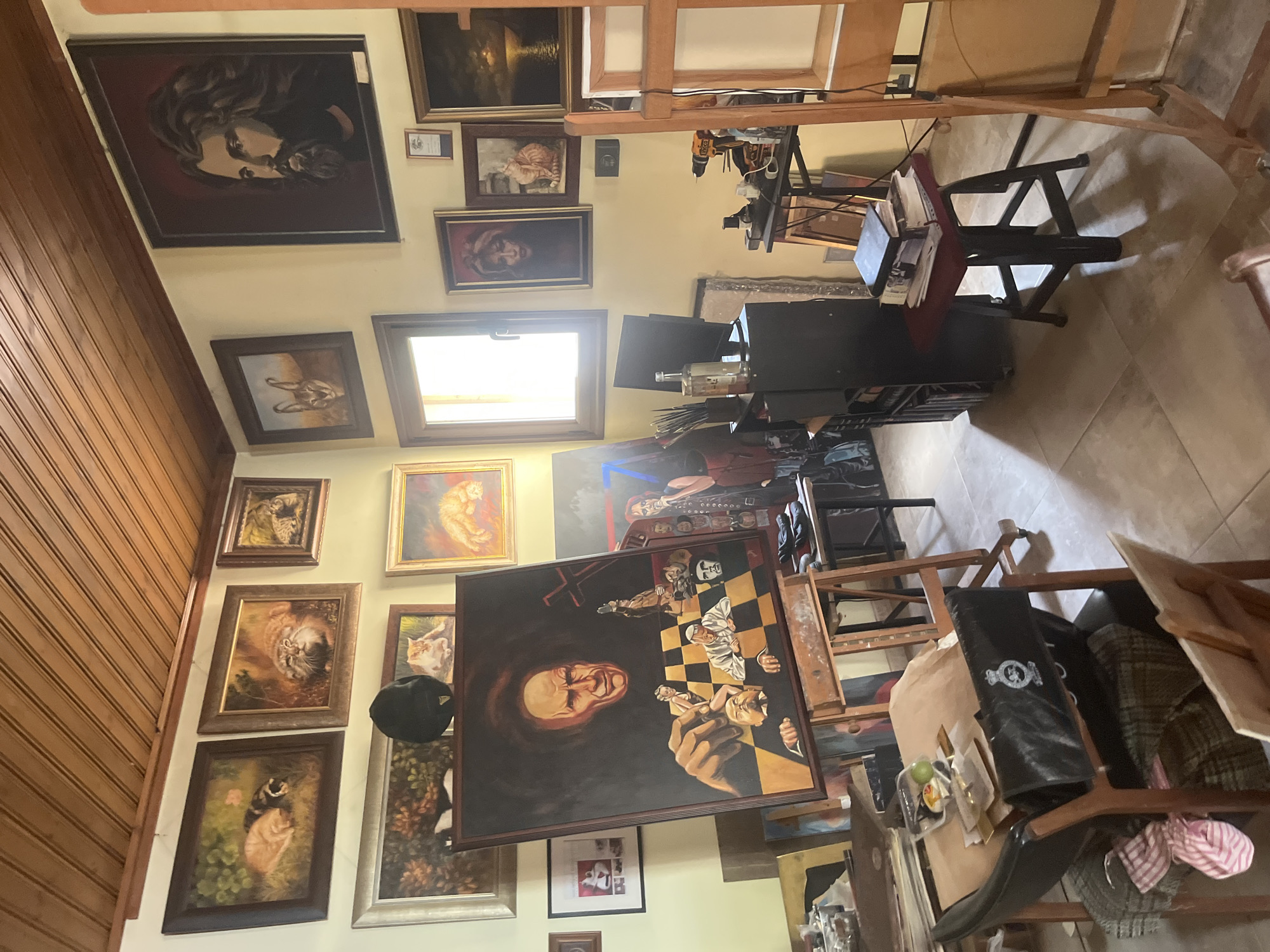 This image depicts the interior of an artist's studio located in the village of Silikou, Cyprus. The room is filled with various paintings, easels, and art supplies, suggesting a working creative space. Dominating the center of the room is a large easel holding a portrait painting of a man's head, rendered in a dramatic style. Surrounding this central piece are numerous other artworks, including portraits of cats, a depiction of a man with long hair and a beard, and abstract or symbolic compositions. The walls and ceiling are paneled with wood, adding a rustic feel to the studio. Art supplies, such as paintbrushes, bottles, and a drill, are visible on tables and shelves, indicating the ongoing nature of artistic endeavors. The lighting suggests it is daytime, with natural light streaming in from a window. The overall impression is one of a cluttered but functional art studio, filled with the artist's creations and tools of the trade.