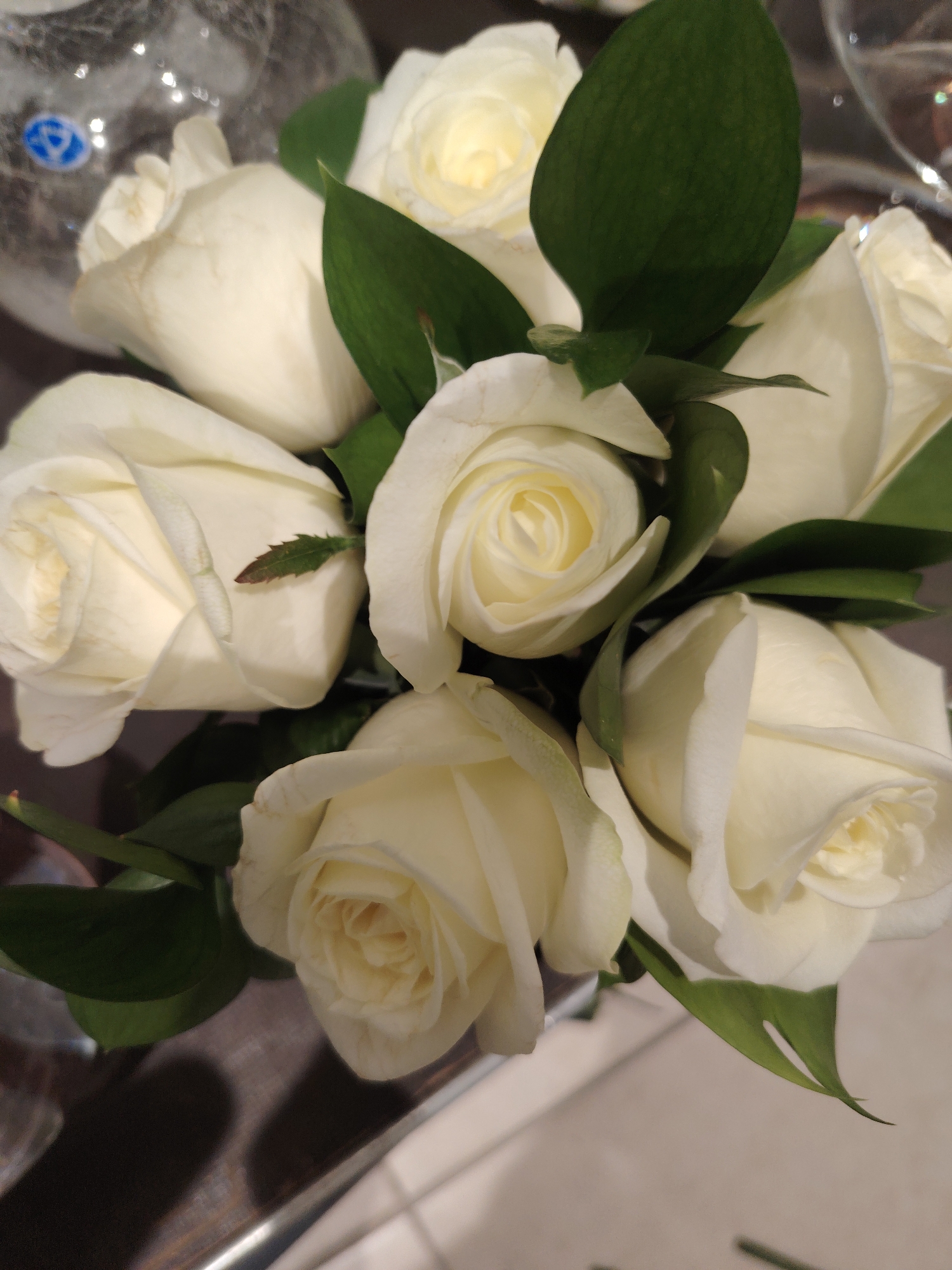 A close-up, vertical shot showcases a bouquet of white roses with green foliage. The roses are arranged in a cluster, with their petals unfurling. Some of the roses are fully open, revealing their intricate centers, while others are still in bud form. The lighting highlights the delicate texture of the petals and the glossy sheen of the leaves. The background is slightly blurred, suggesting a shallow depth of field, and appears to be a domestic setting with a reflective surface and hints of light. The image does not provide any information about the location in the city of Konigil, Uzbekistan, nor does it depict any people or actions.