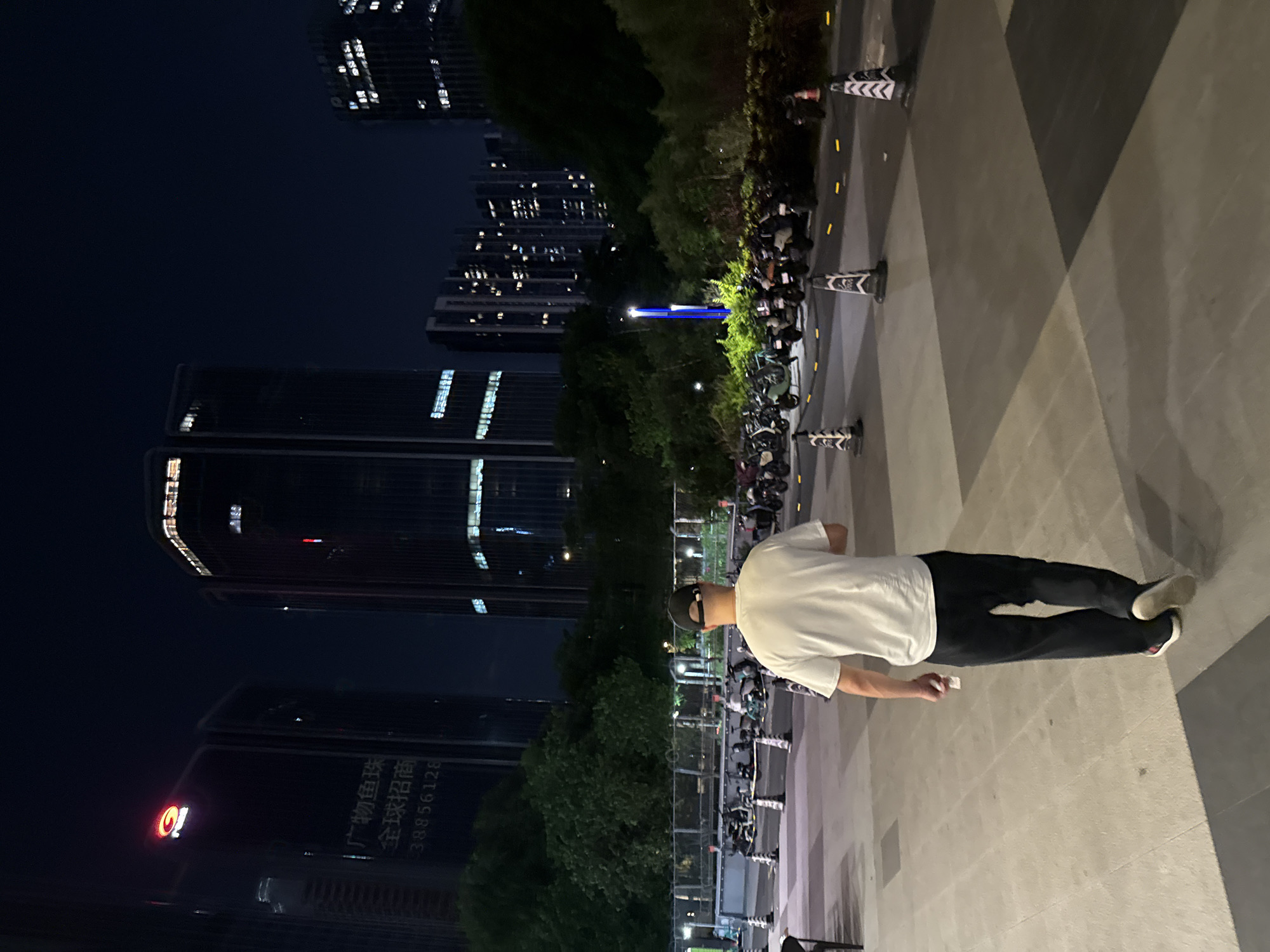 The image depicts a nighttime scene in the Tianhe District of China. In the foreground, a man wearing a white t-shirt and black pants with a black baseball cap walks away from the camera. He is on a paved plaza with large square tiles. In the middle ground, a row of parked scooters or motorcycles is visible, cordoned off by traffic cones. Beyond the parked vehicles, lush green trees form a dense line. In the background, several tall modern buildings are illuminated, with windows lit from within. One building on the left has a red logo and Chinese text. The sky is dark and clear.