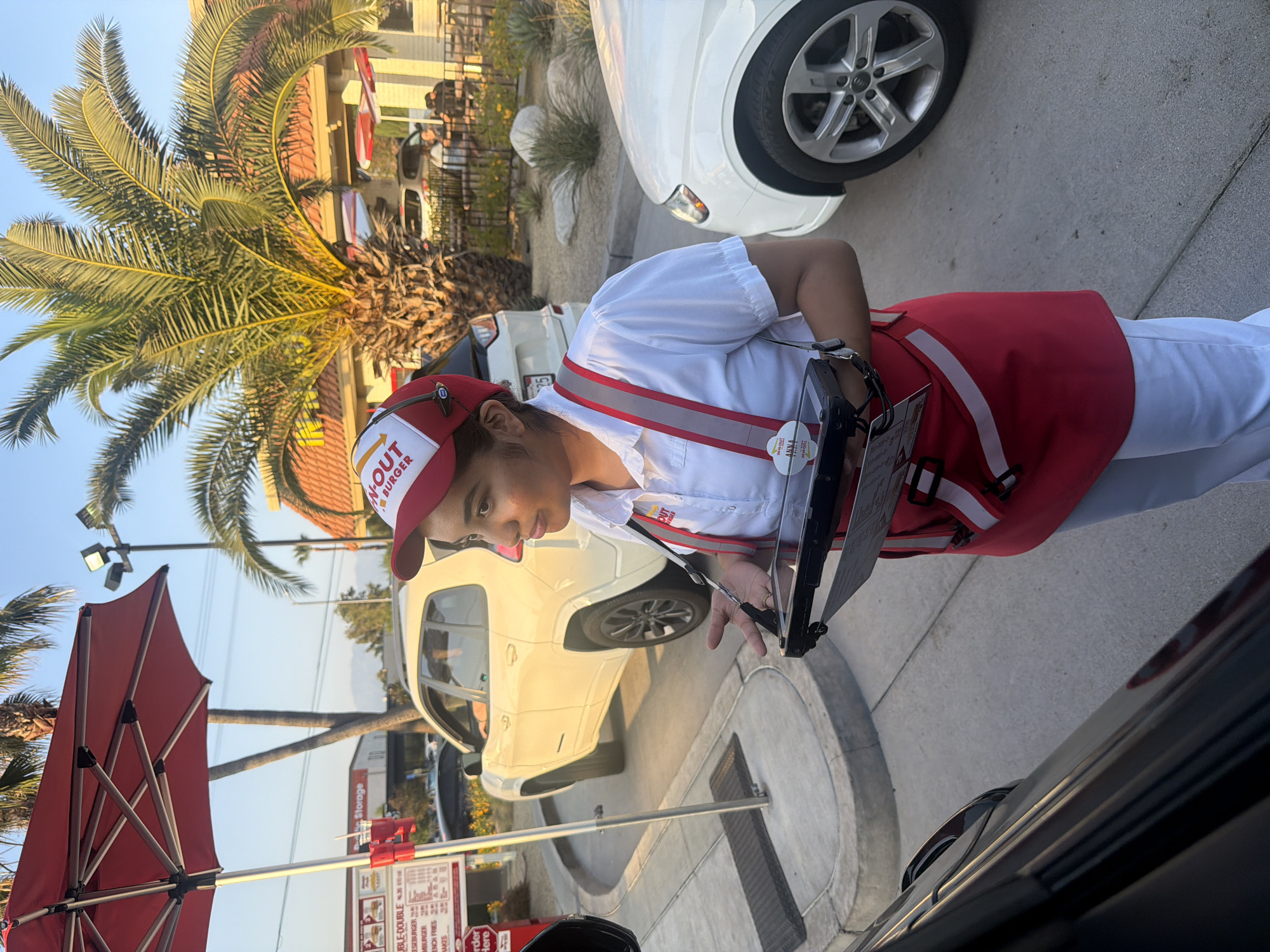 The photograph captures a scene at an In-N-Out Burger drive-through in the city of Los Angeles, USA. A young employee wearing an In-N-Out uniform, consisting of a red and white hat, a white shirt, a red apron with white stripes, and white pants, is taking an order from a customer in a vehicle. She holds an order pad in her hands and is leaning towards the vehicle to communicate with the driver. In the background, other vehicles are visible in the drive-through line, including a white Jeep and a white sedan. A menu board displays food options. A red umbrella provides shade. A palm tree is partially visible in the upper left corner. The sky is a clear blue.