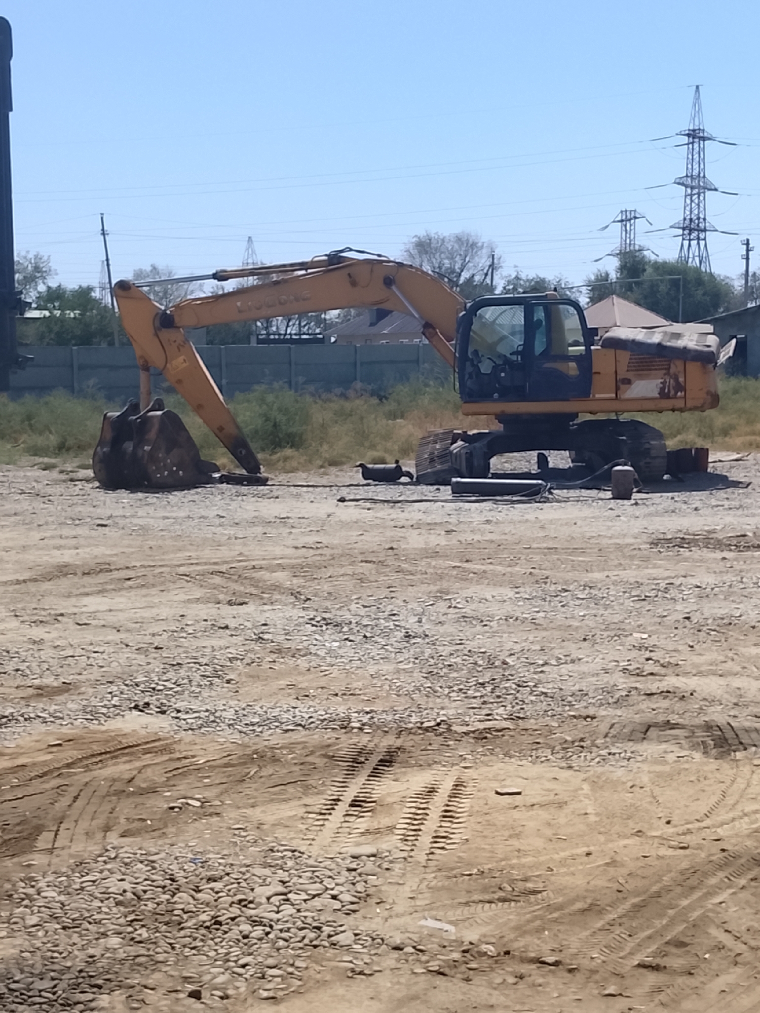 An excavator is parked on a gravel lot in Sabir Rakhimov, Uzbekistan. The excavator is yellow and has a large bucket attached to its arm. The bucket is on the ground in front of the excavator. The excavator is on a construction site, as evidenced by the piles of gravel and the tire tracks in the dirt. In the background, there are some buildings and power lines. The sky is clear and blue.