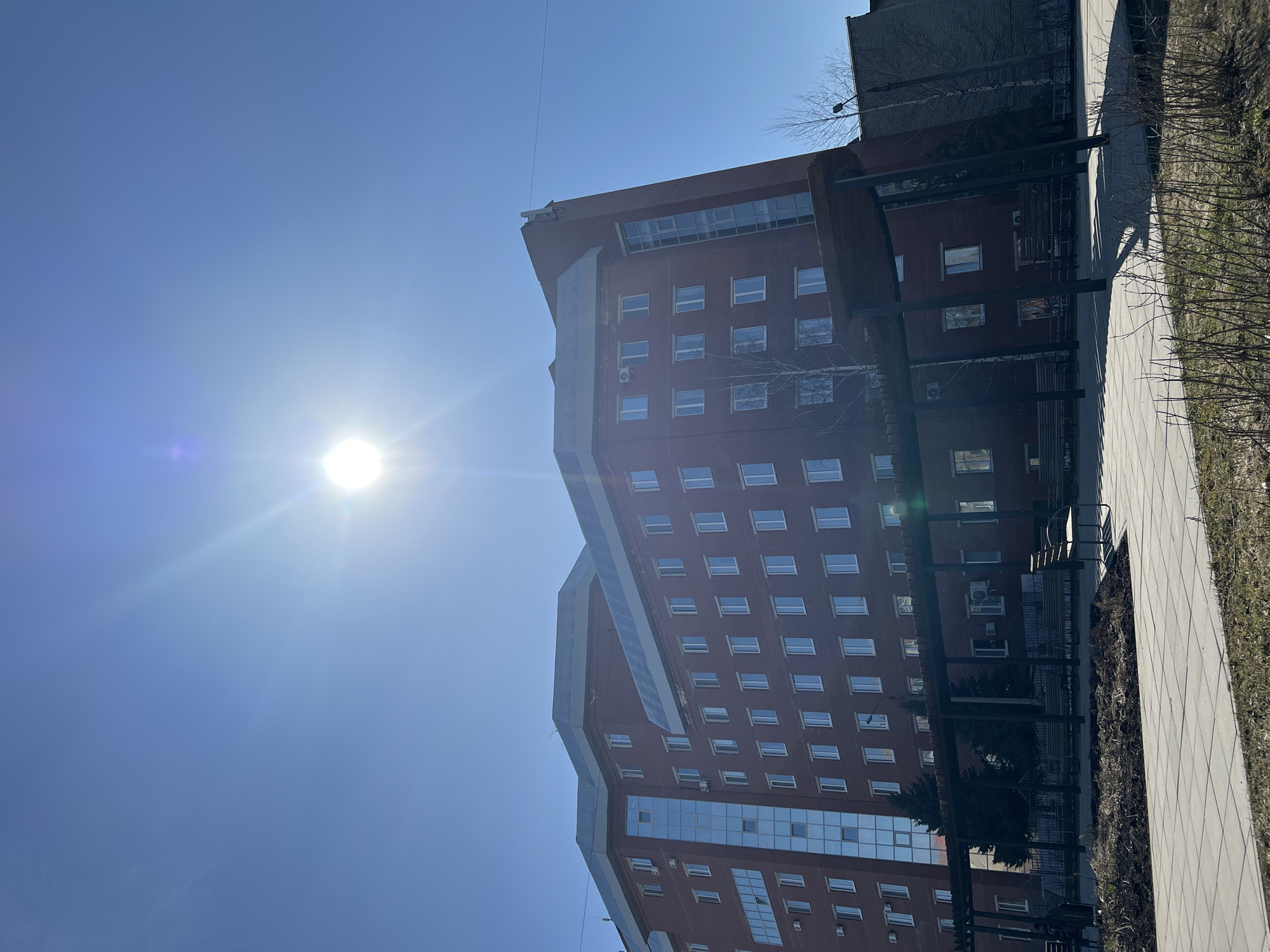 A ground-level, eastward-facing view of a multi-story brick building in Perm, Russia, on a bright, sunny day. The building features numerous windows, a terraced design, and a gray architectural feature. A sidewalk runs along the right side of the frame, bordered by grass and sparse foliage. The sky is a clear blue, with the sun high in the upper-left corner, causing lens flare. An outdoor seating area with a pergola-like structure is visible.