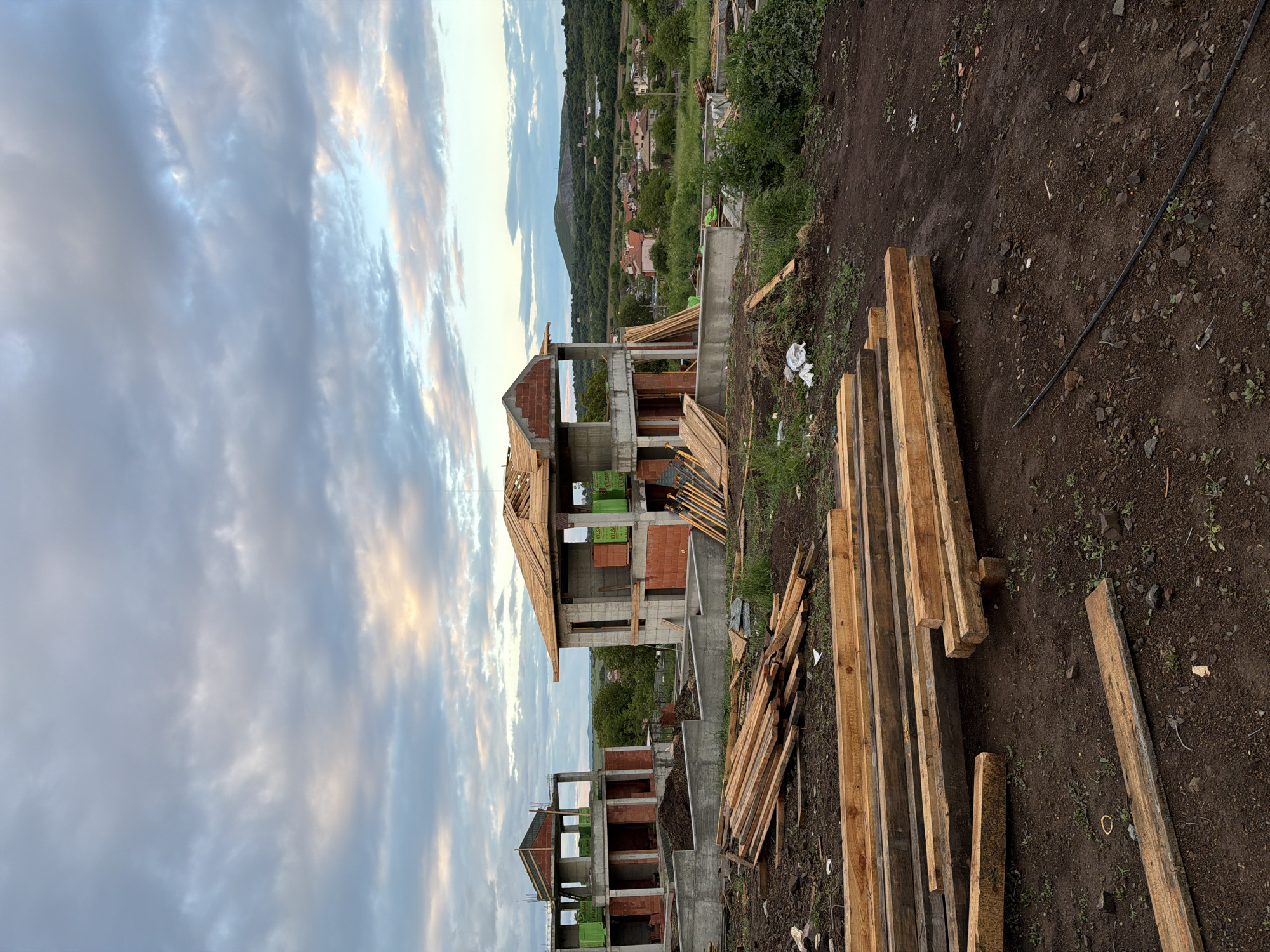 The image shows a construction site in Cherni Vrah, Bulgaria. Partially constructed buildings with red brick walls and concrete supports are visible. Wood planks are stacked on the ground beside the buildings. The sky is cloudy, and there are houses and hills in the background. No people are present. A black hose lies on the dirt.