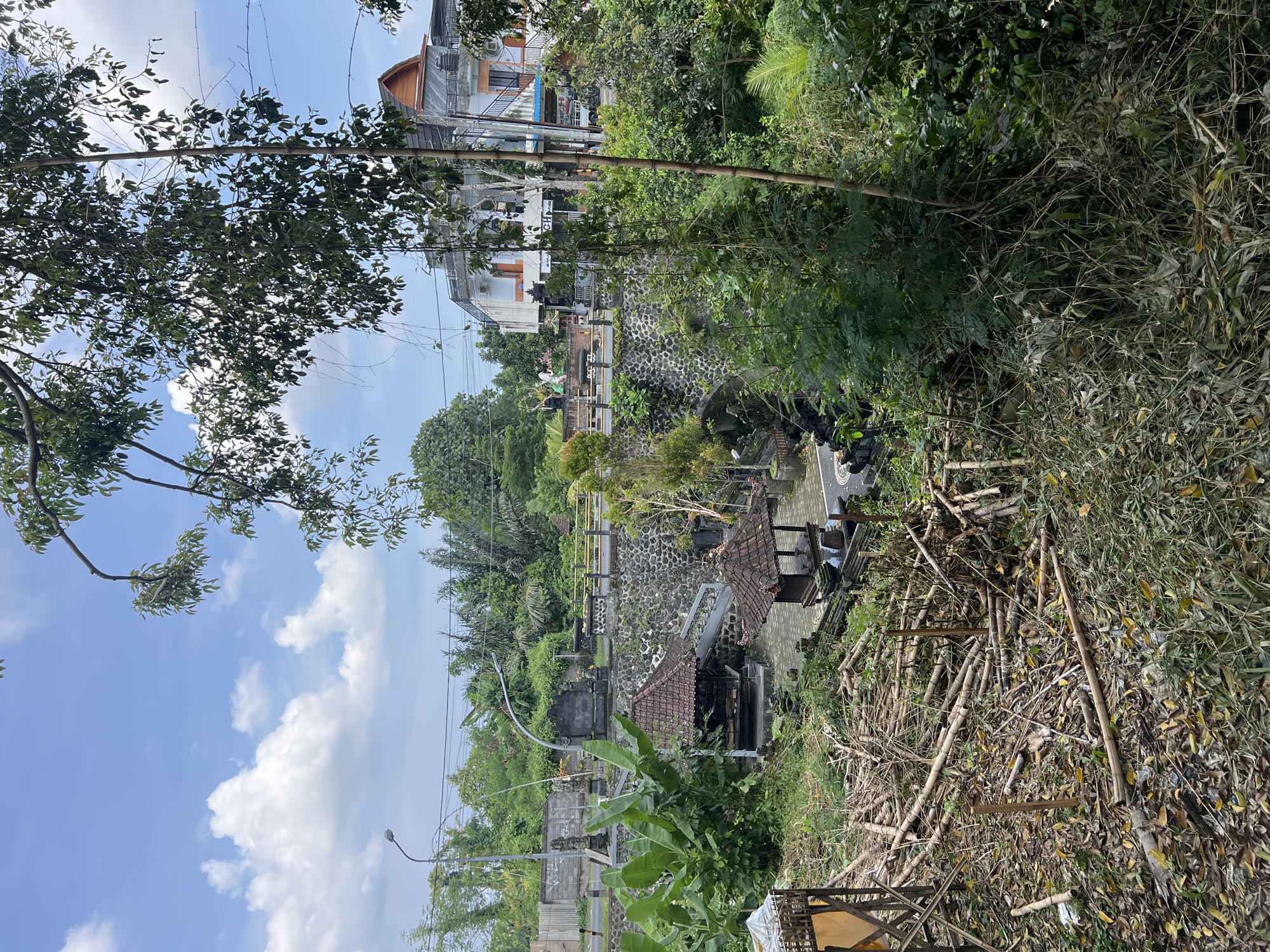 The image depicts a landscape in Buduk, Indonesia, featuring buildings, vegetation, and architectural structures. The sky is visible with scattered clouds. In the middle ground, there are buildings, and further down, structures with tiered levels are visible, constructed with stone and traditional roofing. To the right, there is a pile of dried bamboo. Vegetation is dense throughout the scene. There is an electrical pole on the bottom left.