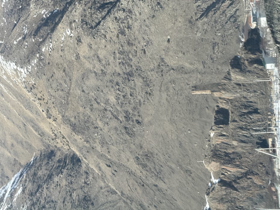 This image presents a wide view of a mountainous landscape in Achkhoti, Georgia, under clear daylight conditions, suggesting a cold season due to visible snow. The upper two-thirds of the frame are occupied by a steep, rugged mountain face, predominantly grey and brown, with minimal vegetation and scattered white patches of snow in depressions and higher areas. Evidence of erosion is visible across the slope.
In the lower third of the image, a tall, weathered stone tower with a rectangular base stands prominently on a rocky outcrop, which also features dark, fractured rock faces. To the left of the tower, on a lower peak of the same outcrop, a white cross is visible. Below the rocky formation and extending into the foreground, a small rural settlement is discernible, consisting of multiple buildings with varying roof colors, interspersed with patches of snow on the ground. Modern infrastructure, including utility poles and overhead power lines, crosses the immediate foreground. No individuals or specific actions are visible within the frame.