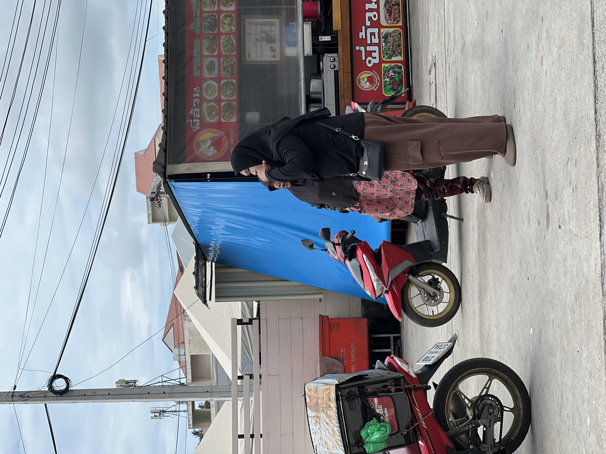 In Ban Bang Rak, Thailand, two women, one in a black hijab and brown trousers, the other in a patterned skirt and darker hijab, stand on a concrete pavement near red scooters. The woman in the foreground is looking upwards and to the right, touching her face. Behind them, a food stall with a red and white sign displays pictures of various dishes. A large blue tarp is visible on the stall. Various wires and a utility pole are present in the background, along with a light-colored structure and buildings with red roofs. One scooter has a cargo box with green bags.