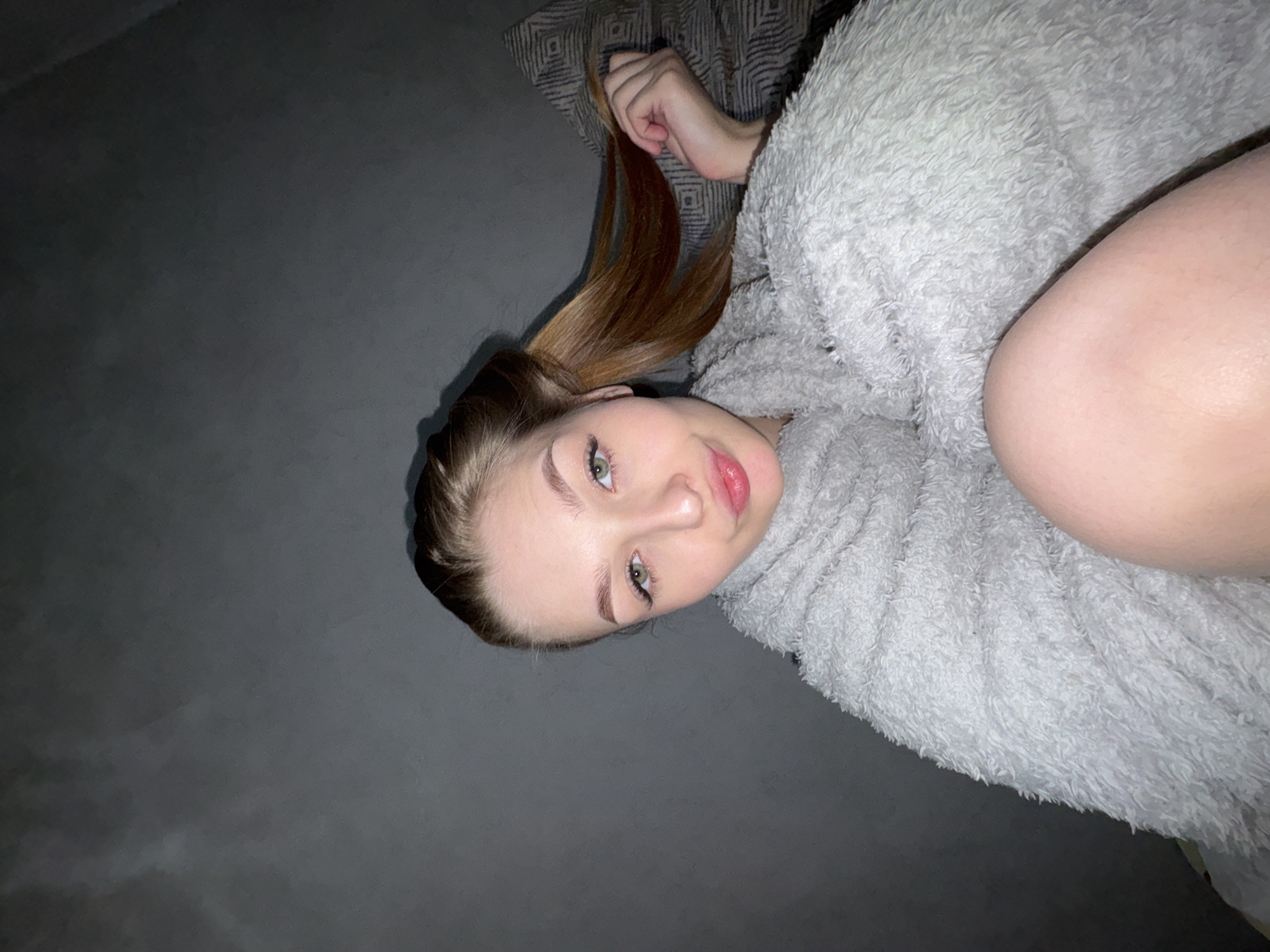 This image, captured in Chaikovsky, Russia, depicts a young woman in an indoor setting, likely a bedroom. She is lying down, looking directly at the camera with a calm, slightly smiling expression. She has light brown hair, which she is holding with her left hand, and striking green eyes framed by dark lashes. Her lips appear full and glossed.
The woman is partially covered by a soft, light grey fluffy blanket or robe, which wraps around her torso. A bare leg or thigh is visible on the right side of the frame, emerging from beneath the blanket. Behind her head, a section of a patterned fabric, possibly a pillow or bedspread, can be seen. The dominant background is a plain, dark grey surface, suggesting a bed or floor. The lighting is dim, indicating it might be evening or nighttime. The overall atmosphere is relaxed and personal. There is no visible text within the image.