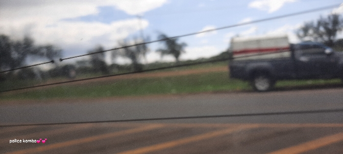 A blurred, wide-angle shot shows a roadside scene with a focus on foreground elements. Utility lines stretch across the frame, partially obscuring the view. A pickup truck with a camper shell is parked on the right side of the road. Beyond the road, a field of green grass leads to distant trees under a cloudy sky. The overall image quality is poor due to the blurriness. "police komba" and a heart emoji are added in the bottom left corner.
