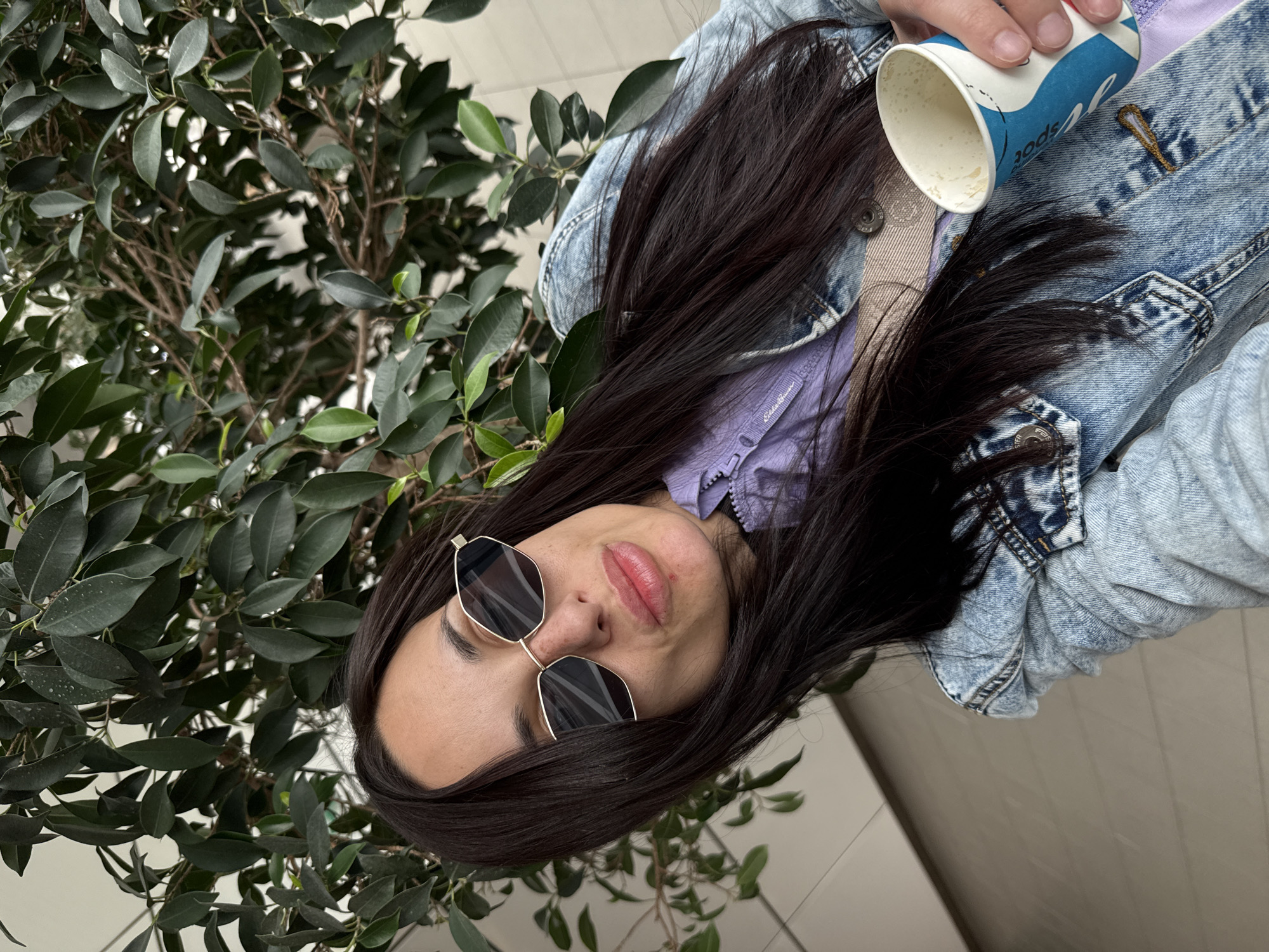 A young woman with dark hair and sunglasses stands next to a leafy tree, possibly indoors, holding a blue and white paper cup. She wears a light blue denim jacket over a purple garment. The background shows a neutral-toned floor and wall, suggesting an interior space. While the image does not contain explicit landmarks, the setting could be an interior space of a commercial building in any city and the specific location in Sofia cannot be determined from this image alone.