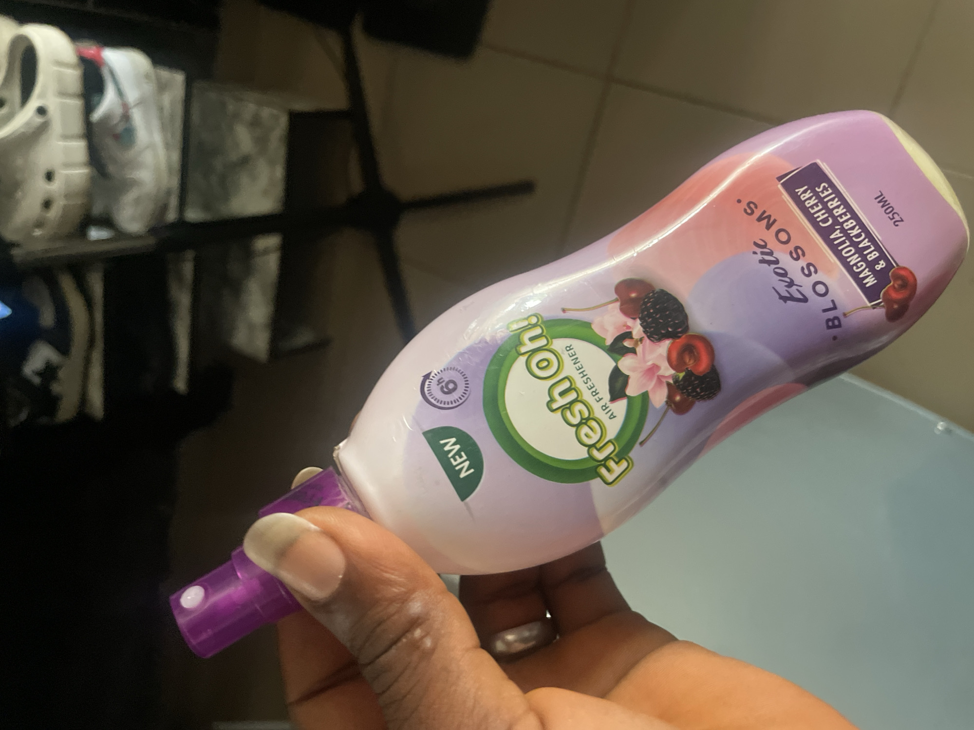 A person's hand with a ring on their left hand holds a purple-capped spray bottle of "Fresh Oh! Air Freshener" in the "Exotic Blossoms" scent, featuring "Magnolia, Cherry & Blackberries." The bottle has a pastel gradient design. The background is blurred, showing shelves with shoes. The image is taken indoors.