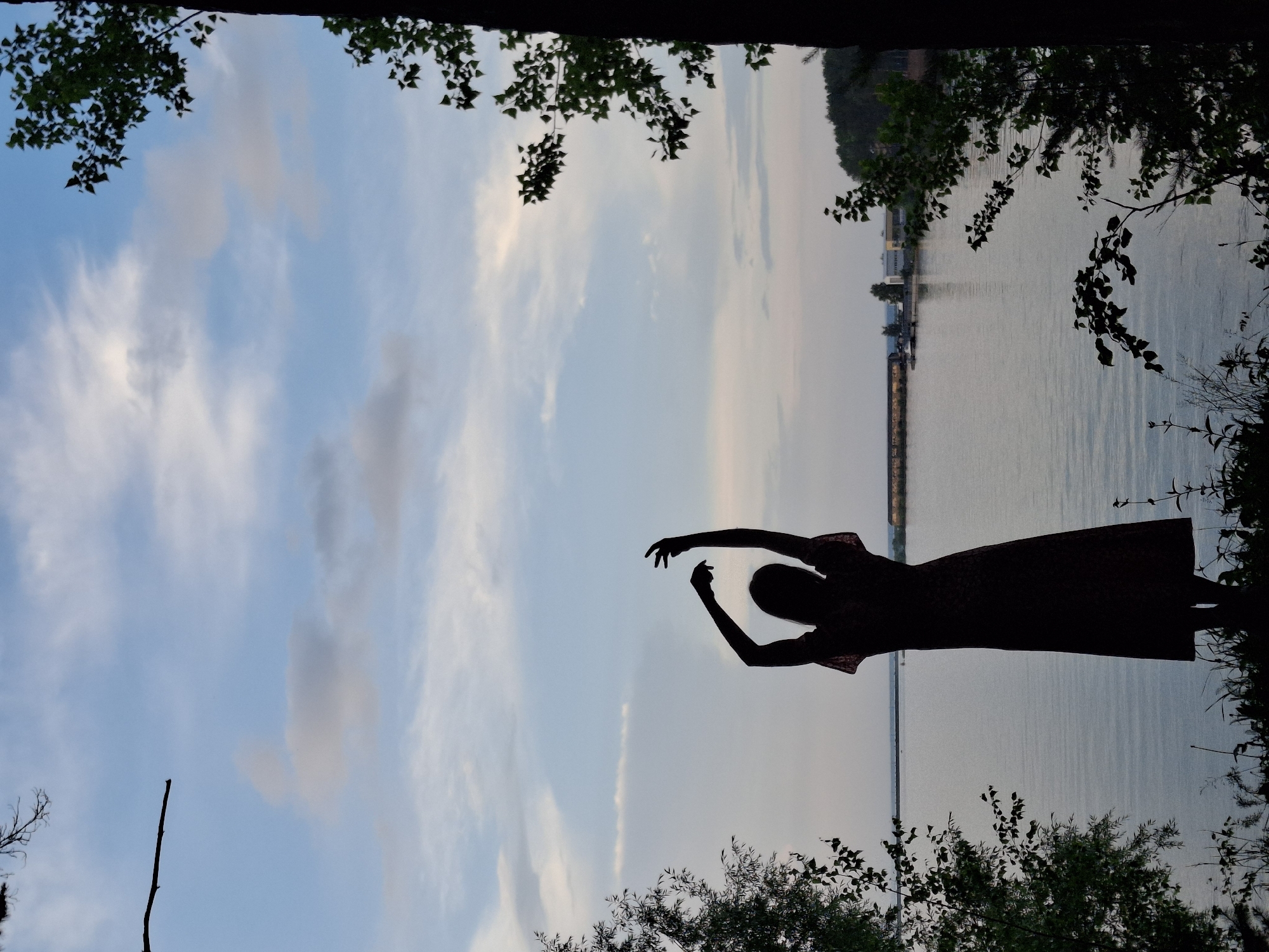 A silhouetted figure of a woman in a long dress stands facing a body of water, with her arms raised and hands in a graceful pose. The scene is framed by dark trees on the left and right, with branches and leaves visible in silhouette. The sky above is a soft blue with scattered white and gray clouds. In the distance, across the water, there are buildings and a line of trees. The water is calm and reflects the sky. The foreground shows dark vegetation and grass. The image captures a tranquil moment by the water, possibly during dusk or dawn, given the soft lighting and muted colors. The location is the Kama River in the city of Chaikovsky, Russia.