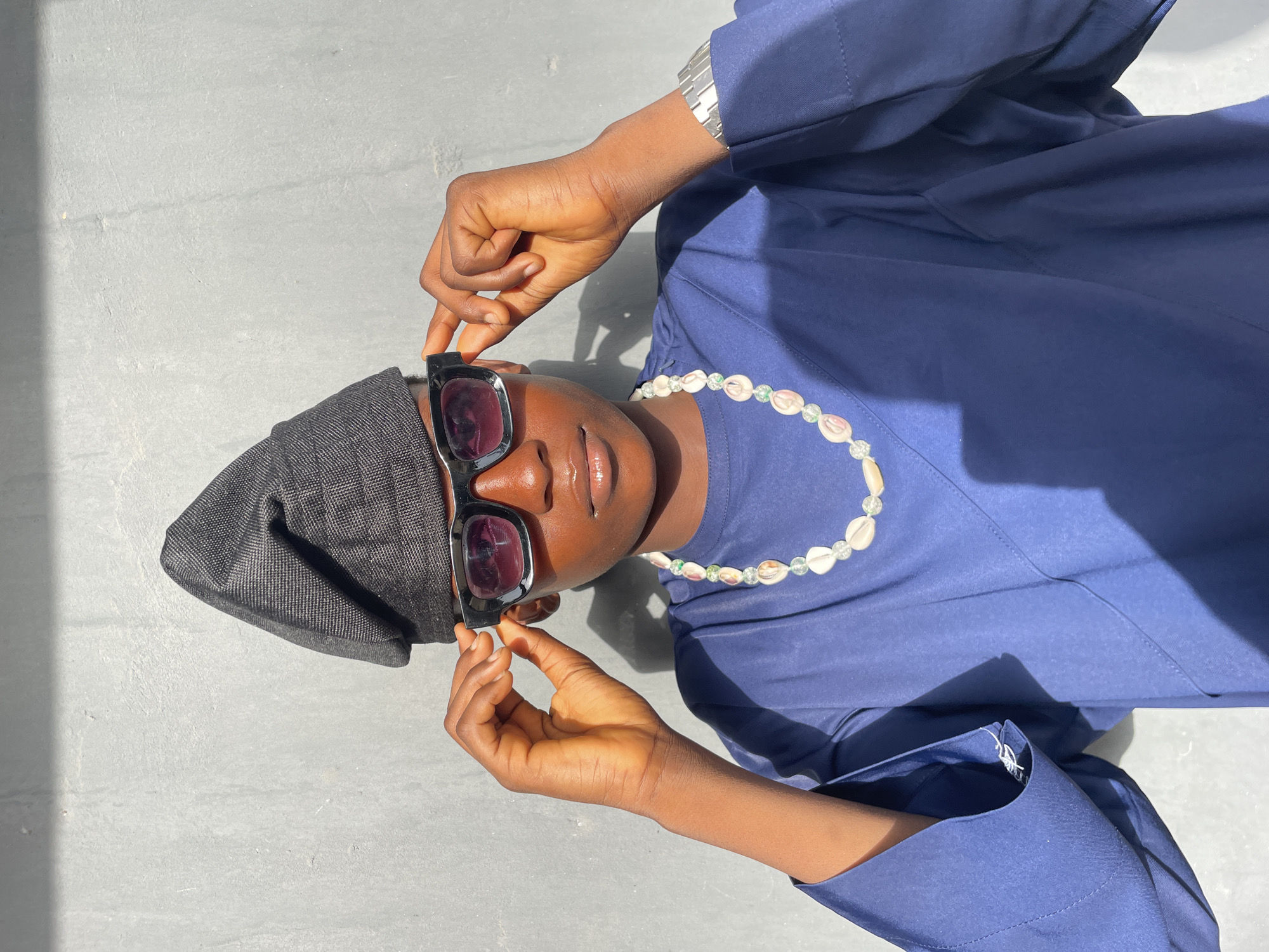 This portrait photograph, set in Badagry, Nigeria, features an individual with a dark skin tone posing outdoors under bright daylight. The subject is captured from an overhead perspective, lying or leaning back on a light grey, textured surface, possibly concrete. They are dressed in a navy blue traditional garment, potentially a kaftan, and a dark grey or black cap that resembles a traditional Fila. Black, square-framed sunglasses with dark lenses are worn on their face, with both hands positioned to touch or adjust the frames near the temples. A necklace crafted from small, light-colored shells interspersed with clear and light green beads is visible around their neck. A silver-toned watch or bracelet is worn on their left wrist. Distinct, sharp shadows are cast on the left side of the frame, indicating strong, direct sunlight. The person's expression is neutral and composed, with slightly parted lips, conveying a sense of relaxed style within a cultural context. No visible text is present in the image.