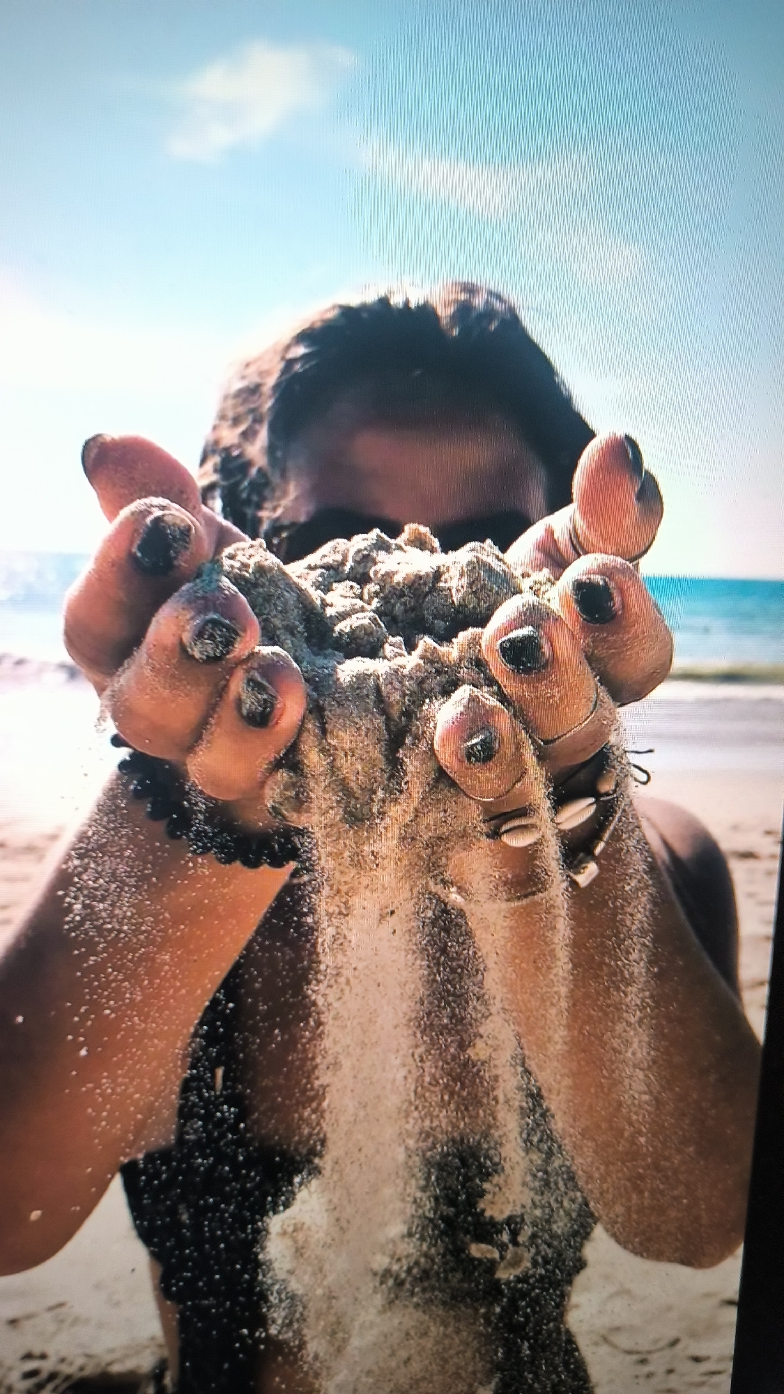 The image shows a person's hands holding a pile of sand, with the sand falling through their fingers. The person appears to be on a beach, with the ocean and a clear sky visible in the background. The lighting suggests it is daytime, possibly mid-morning or afternoon. The person has dark hair and dark nail polish. They are wearing some jewelry, including bracelets and rings. The overall mood of the image is serene and peaceful, with a focus on the natural elements. There is no visible text in the image. The location is not identifiable from the image alone, and the provided location context of Kovrov, Russia is not evident from the visual information.