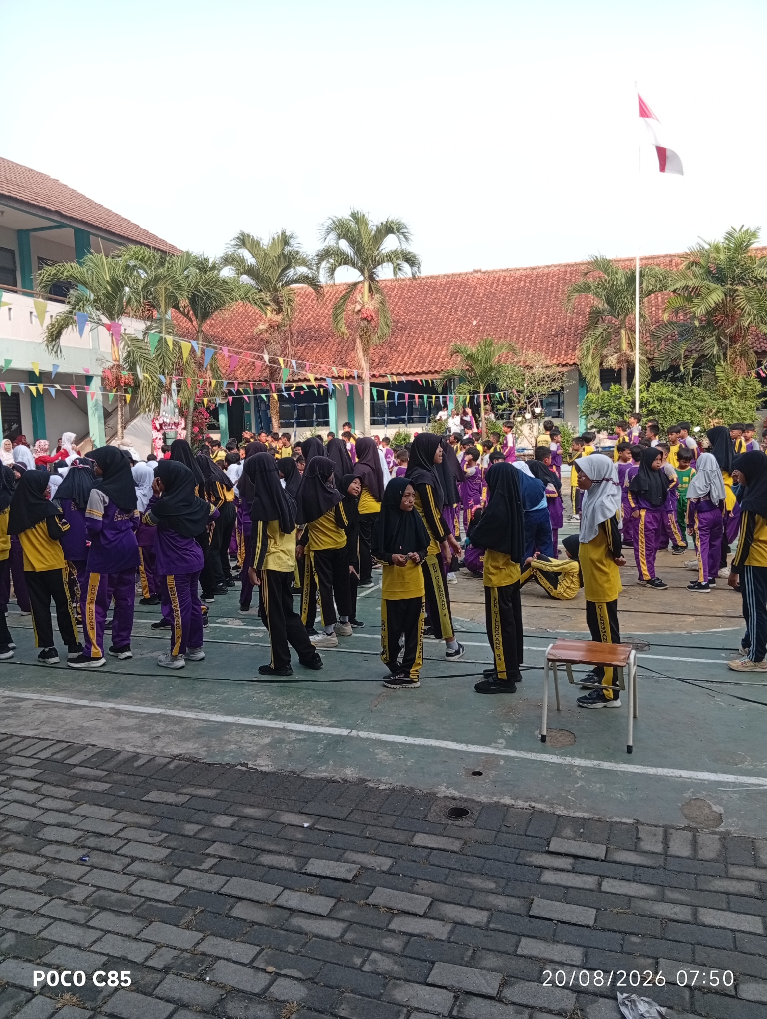 A group of students, predominantly girls wearing purple and yellow uniforms with headscarves, are gathered in an outdoor courtyard. Some are standing in formation, while others appear to be engaged in an activity. The environment includes buildings with red tiled roofs, palm trees, and colorful triangular flags strung across the area, suggesting a school event. A flag pole with an Indonesian flag is visible in the background. The ground is a combination of paved brick and a green court surface with white lines. A small wooden stool is present in the foreground. The timestamp "20/08/2026 07:50" and "POCO C85" are superimposed on the image. The scene is likely at a school in South Tangerang, Indonesia, given the context provided by the timestamp and potential school setting.
