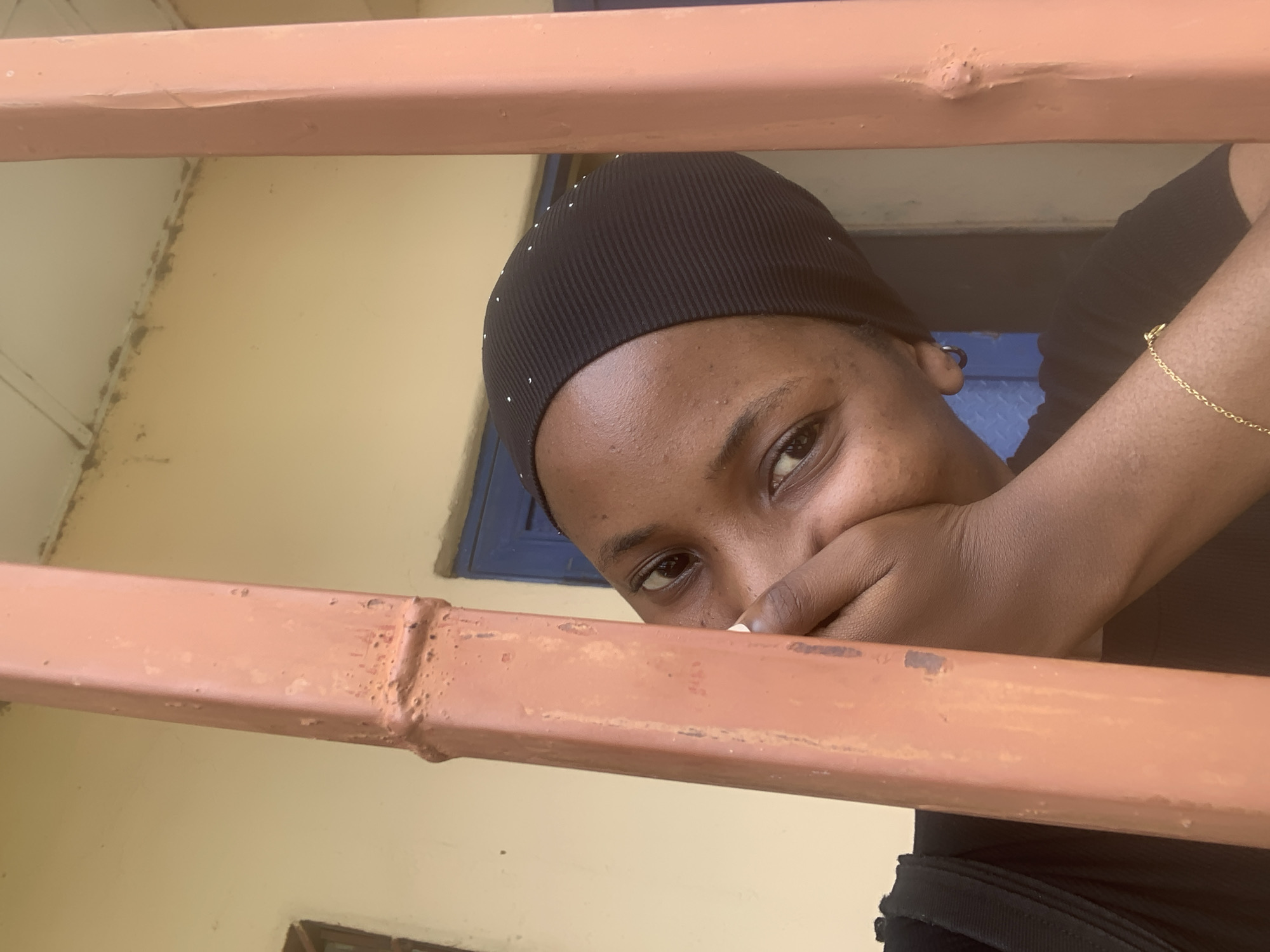 This is a close-up, medium shot of a young Black woman looking at the camera from behind a reddish-orange metal bar. She is wearing a black headscarf with small white embellishments. Her face is turned to the side, and her right hand is covering her mouth, with only her eyes and a bit of her nose and chin visible. She has dark brown eyes and dark skin. A thin gold chain bracelet is visible on her left wrist. She is wearing a black top.
The background is a light yellow wall with a window that has a blue frame. The metal bar in the foreground appears to be part of a railing or fence. The lighting suggests it is daytime. The scene appears to be outdoors, possibly in an urban or semi-urban setting, given the appearance of the wall and window. The woman's expression is somewhat coy or playful, with her eyes making direct contact with the viewer. There is no visible text in the image. The context provided indicates this image was taken on Rukuba Road, Nigeria.