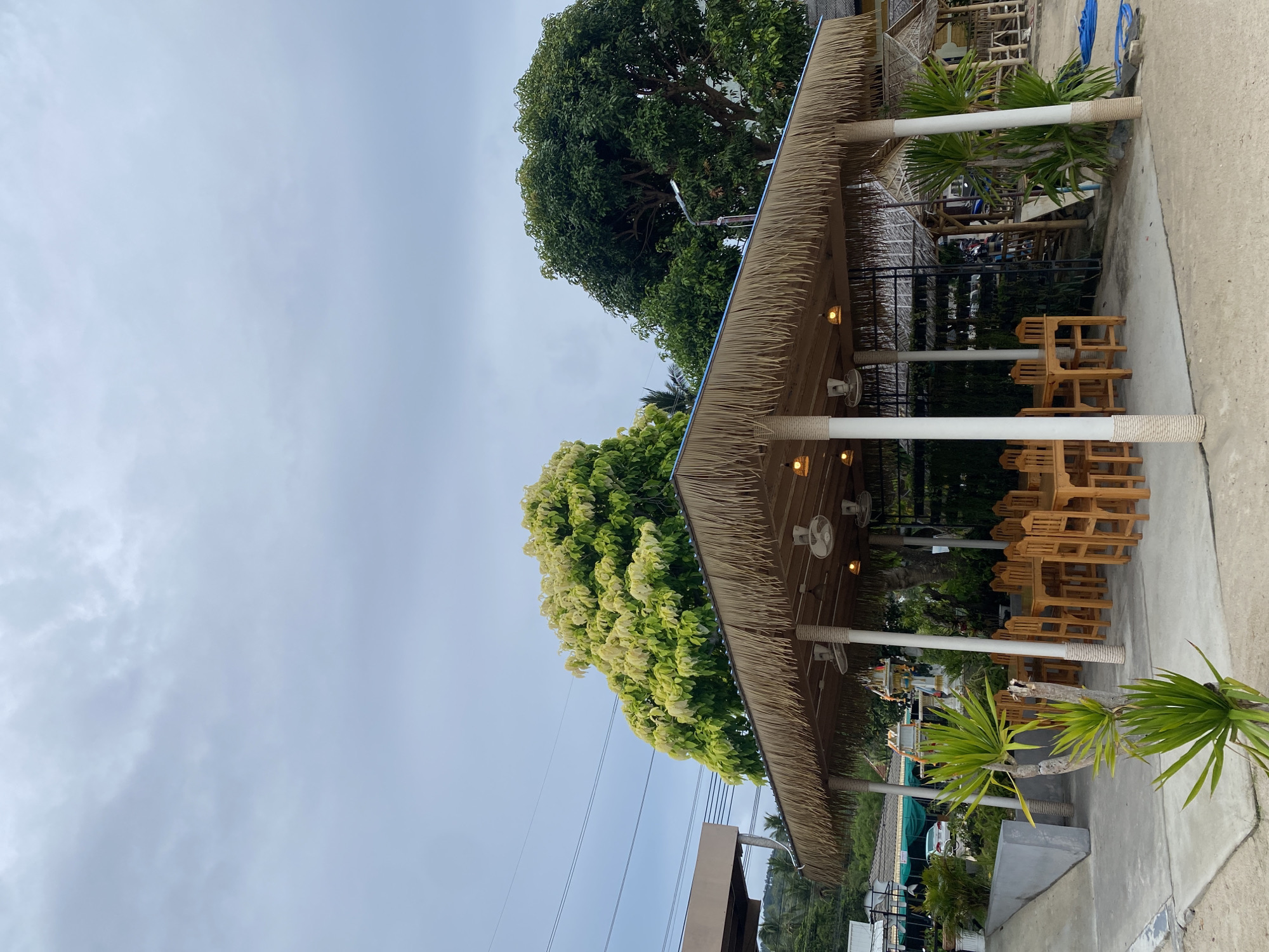 The image shows an outdoor dining area, likely a restaurant or cafe, in Baan Thong Krut, Thailand. The dining area is covered by a thatched roof supported by white pillars adorned with rope. Several wooden chairs are neatly arranged around tables. Ceiling fans are visible under the roof. The surrounding environment includes trees, a concrete surface, and glimpses of other structures and vegetation, suggesting an outdoor setting. The sky is overcast.