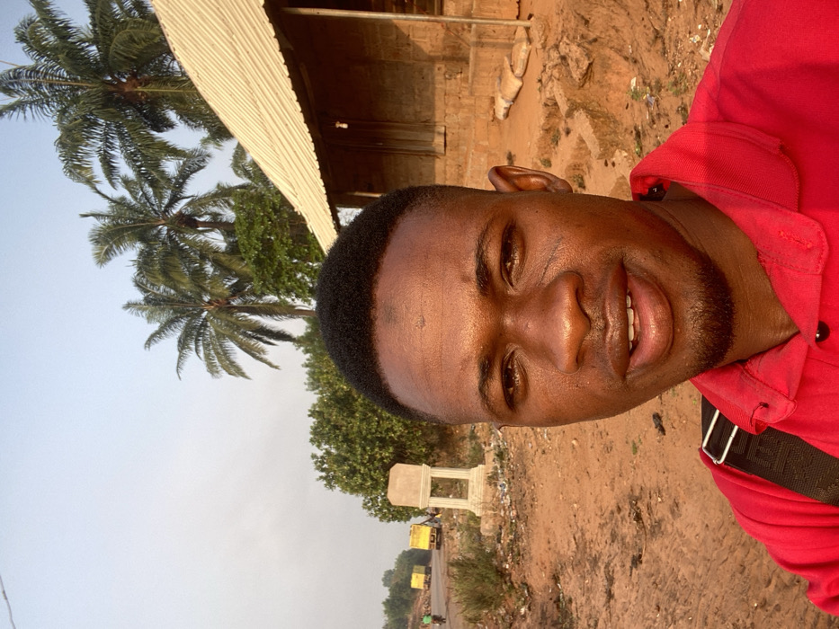A man wearing a red shirt smiles into the camera. He is outside on a dirt road next to trees and some buildings. The sky is light blue and there are trucks in the distance. The photo is slightly blurry and the man's skin is shiny.
