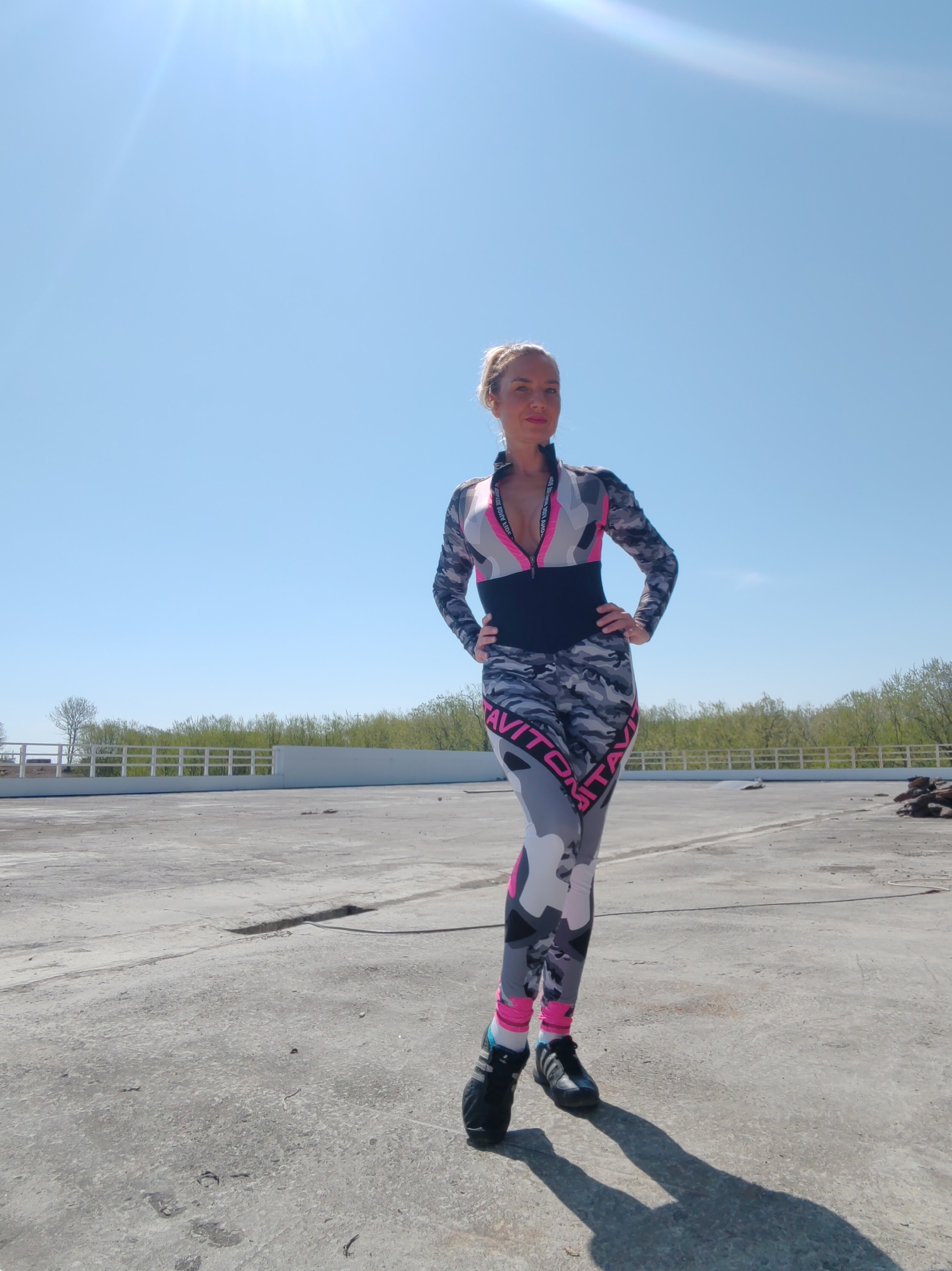 The image depicts a female individual standing outdoors on a large, weathered concrete surface, likely a rooftop or an expansive paved area, in Petropavlovsk-Kamchatsky, Russia, under a clear blue sky.
The woman, positioned slightly off-center to the left, is an adult with blond hair pulled back into a ponytail. She wears a form-fitting, full-body athletic suit featuring a grey, black, white, and hot pink camouflage pattern. The suit has long sleeves and full-length legs, with a distinct black horizontal band cinching the waist. The chest area has a visible zipper, partially open, revealing an inner black layer or neckline with white text that appears to read "GRAVITY ON". Similar "GRAVITY ON" text is printed vertically along the outer side of both legs in hot pink. She wears dark athletic shoes, primarily black with light blue and white accents, and white socks. Her hands are placed on her hips, and she looks directly at the camera with a subtle smile. Strong sunlight from the upper left casts a distinct shadow of her figure to her right.
The concrete ground displays various cracks, stains, and scattered small debris, indicating wear and exposure. In the mid-ground, a white, slatted fence or barrier extends horizontally across the frame, acting as a demarcation. Behind this fence, a line of trees is visible, with both green foliage and bare branches, suggesting an early spring or late autumn season. To the far right on the concrete surface, a dark, irregular pile of material, possibly rubble or wood, is present. The upper half of the image is dominated by an uninterrupted, bright blue sky with a distinct sun flare originating from the upper-left quadrant.