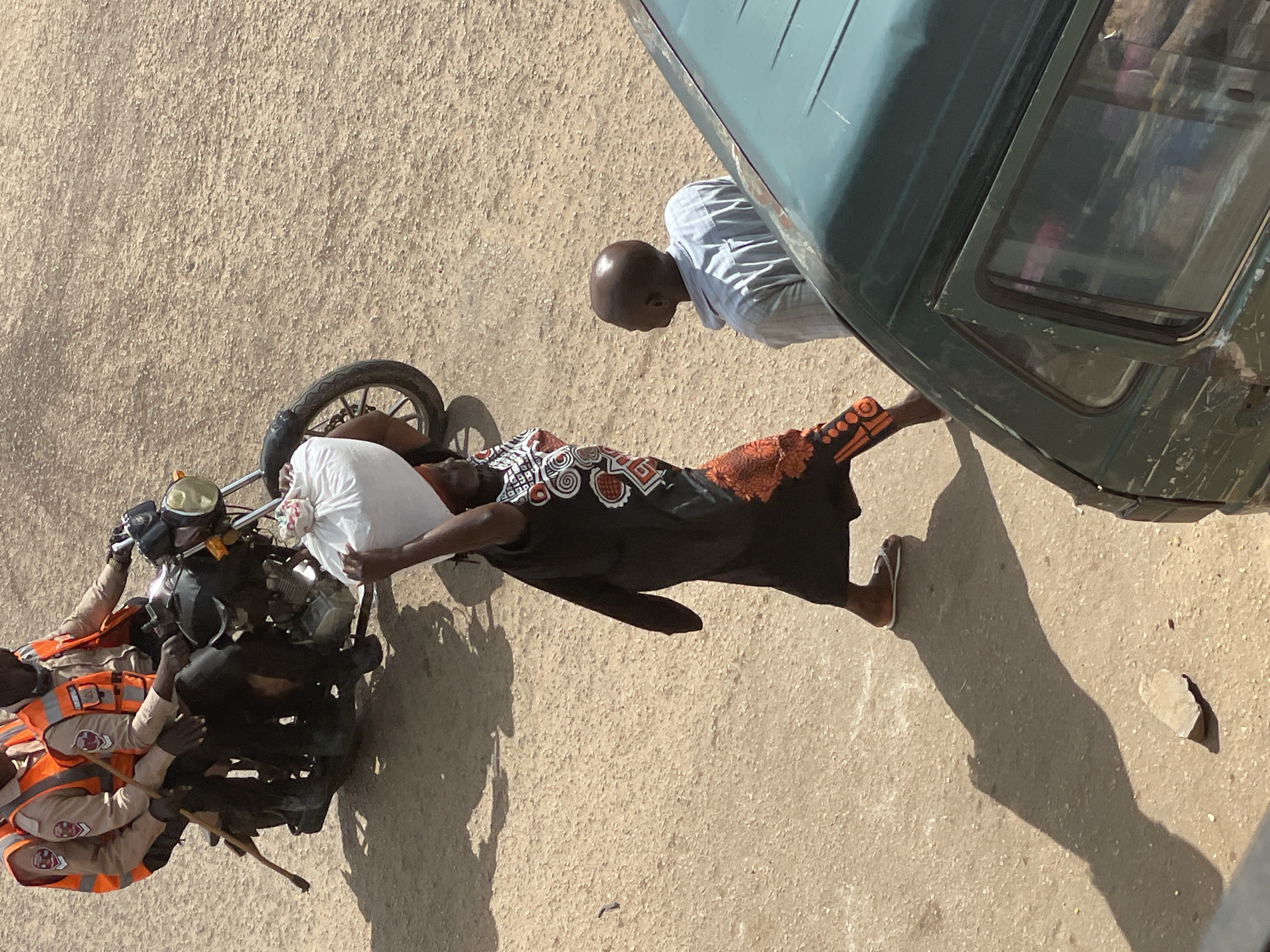 In a dusty, outdoor urban environment, three individuals are visible. Two individuals, wearing orange safety vests over tan shirts and black pants, are seated on a dark-colored motorcycle. They appear to be law enforcement or security personnel, with one holding a wooden stick. In front of the motorcycle, a third individual, wearing a dark patterned dress with orange accents and flip-flops, carries a large, white bag on their head. To the right of this individual, a bald person wearing a light blue pinstripe shirt stands looking to their left. A portion of a dark green vehicle is visible in the foreground, and the ground is a dry, sandy surface. The lighting suggests a bright, sunny day, casting long shadows.