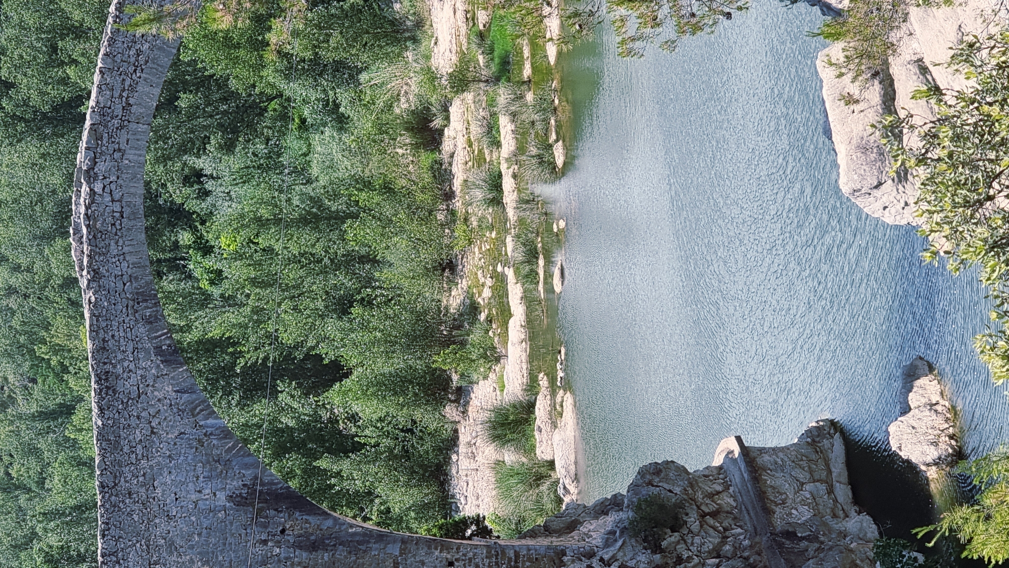 An aged stone bridge spans a body of water, with lush green trees and vegetation covering the background. The bridge, constructed of grey stones, exhibits an arched shape and appears weathered with some stones missing from its top. The water below is calm, reflecting the surrounding greenery and sky, with scattered rocks and patches of grass emerging from its surface near the edges. On the left side of the frame, a rocky cliff face descends to the water, with a narrow stone pathway or ledge built into its side. The scene is set in a natural environment, likely a rural or park area, given the abundant foliage and the rustic nature of the bridge. The overall impression is one of natural beauty and historical architecture. This scene is located in the city of Tortellà, Spain.