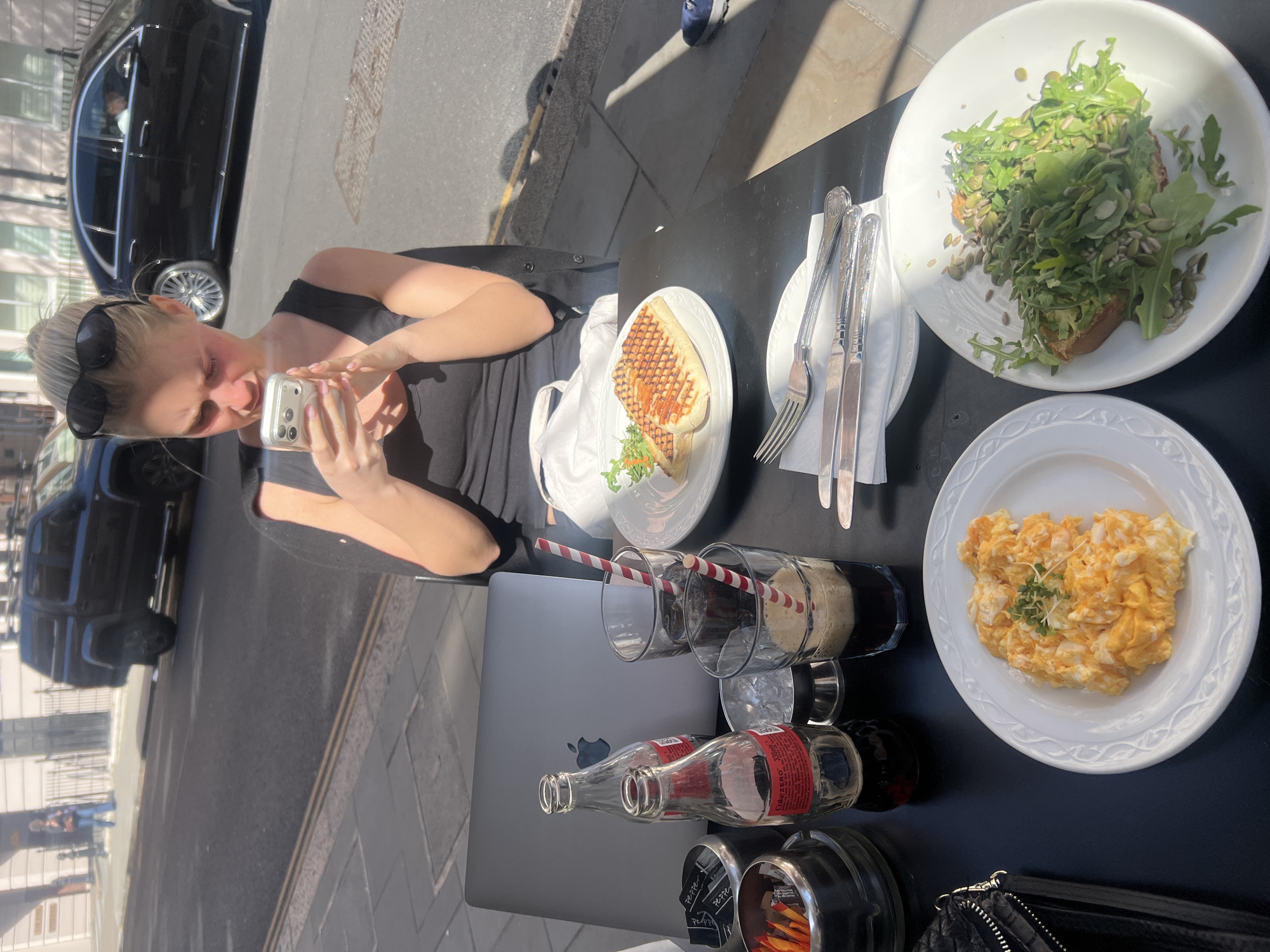 This media file captures an outdoor cafe scene in London, United Kingdom, on a bright and sunny day. The primary subject is a blonde woman, with her hair pulled back and sunglasses resting on her head, wearing a black top. She is focused on taking a picture of the food on the table with a silver smartphone. Her face shows some redness, possibly from sun exposure.
The black outdoor cafe table is laden with various dishes and drinks. On white plates, there's a serving of scrambled eggs garnished with microgreens, a slice of avocado toast heavily topped with arugula and pumpkin seeds, and a grilled panini or sandwich accompanied by a small portion of arugula. Beverages include two tall glasses with red and white striped straws containing dark liquids, and an empty glass bottle of Coca-Cola Zero Sugar (200ml) with its distinctive red label.
Other items on the table include a closed silver Apple MacBook laptop, a black quilted handbag with a silver zipper, and cutlery (forks and knives) resting on a white napkin. A dispenser holding sugar packets, partially labeled "PEPPER," is also visible.
The background reveals a typical city street with strong sunlight creating pronounced shadows. Parked black vehicles, including a sedan and an SUV, are visible on the street, alongside buildings reflected in their windows. The clear skies and bright illumination suggest it is daytime.