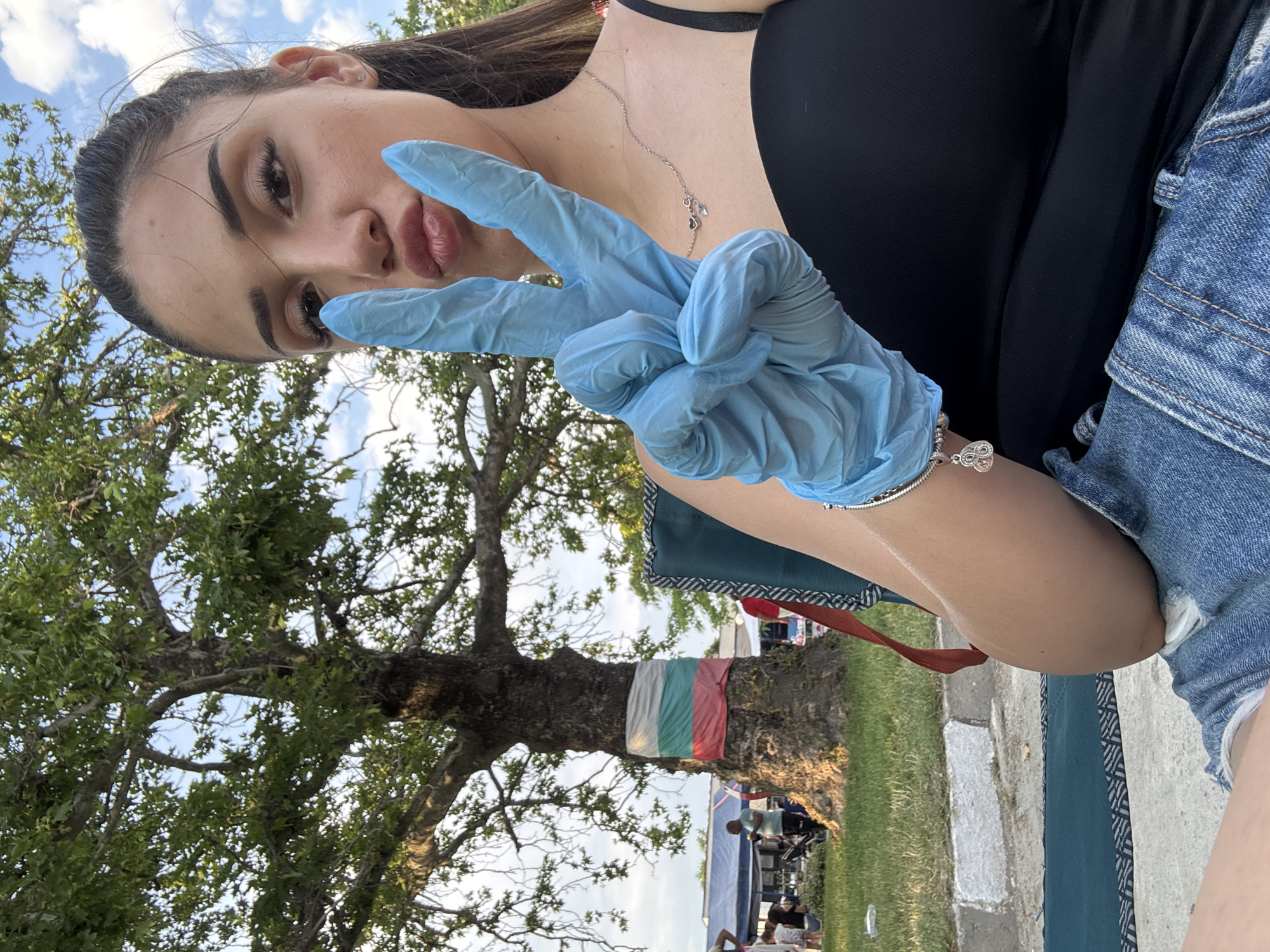 A young woman is prominently featured in the foreground of an outdoor scene in Kostievo, Bulgaria. She is seated, facing the viewer, with her head slightly tilted and her lips pursed in a pout. Her left hand, encased in a light blue disposable glove, is raised, forming a "peace sign" gesture. She has long, dark hair pulled back, and her facial features include dark eyebrows and visible makeup. She wears a black spaghetti-strap top, denim shorts, a silver necklace with two small heart pendants, and a silver charm bracelet on her left wrist. She is seated in a dark green folding chair with a geometric pattern.
In the background, a large, mature tree with dense green foliage occupies the left side of the frame. Wrapped around the lower trunk of this tree is a fabric band displaying the horizontal stripes of white, green, and red, consistent with the flag of Bulgaria. To the left and behind the tree, an outdoor gathering is visible, featuring multiple individuals under blue and white canopies or tents. These individuals are engaged in various activities, some standing and others seated. The ground consists of green grass and a light-colored paved curb. The sky is bright blue with scattered white clouds, indicating daytime conditions.