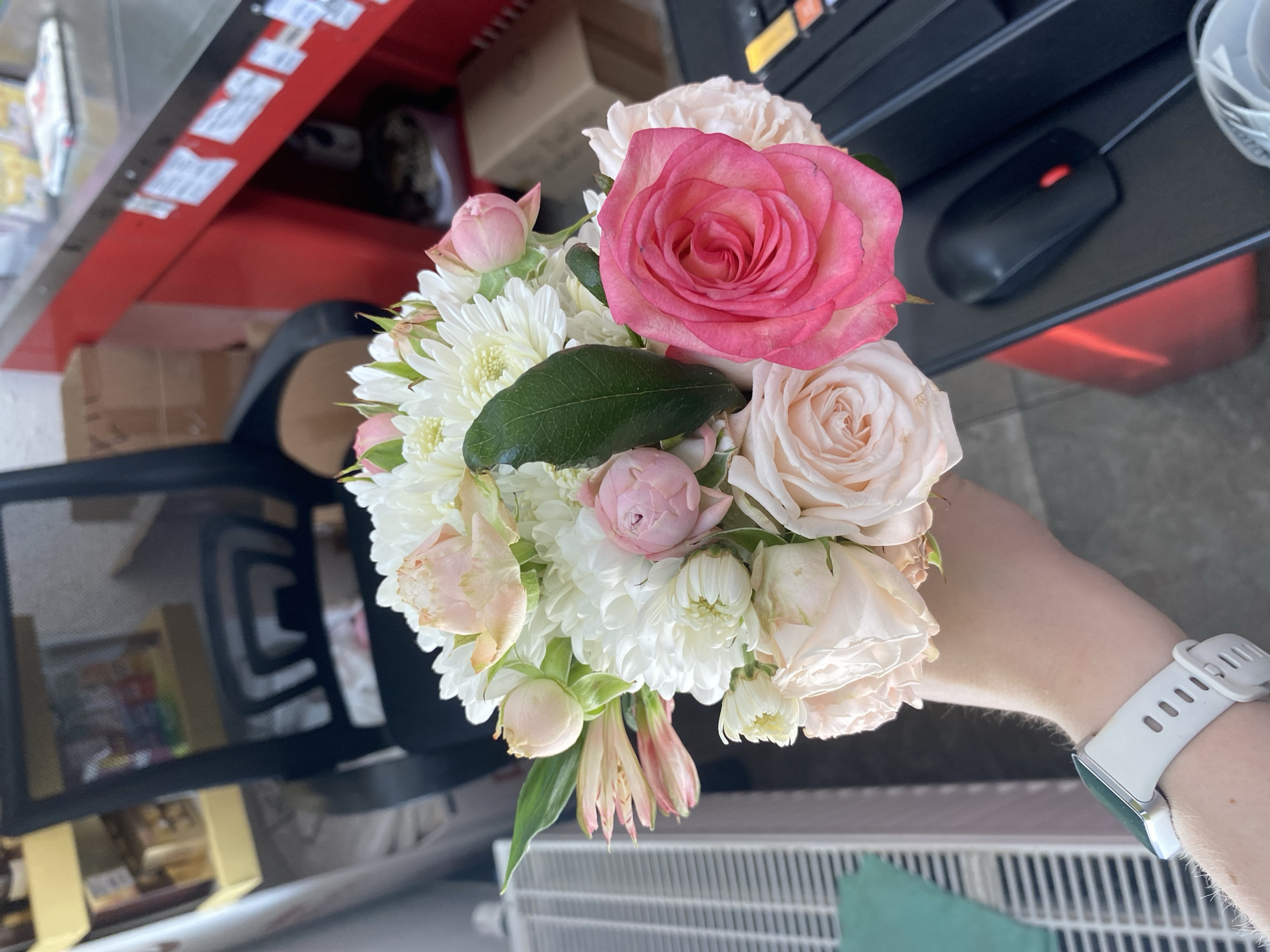 A high-angle shot depicts a hand holding a bouquet of flowers. The bouquet is composed primarily of white chrysanthemums, peach-colored roses, and a single bright pink rose as the focal point. A solitary green leaf is visible among the blooms. The hand wearing a white smart watch is holding the bouquet against a backdrop of office supplies. The background includes a black office chair with a mesh back, a red shelf stocked with various items, and a black computer mouse on a dark surface. Below the bouquet, a white radiator is partially visible. The location is likely an office in Yekaterinburg, Russia.
