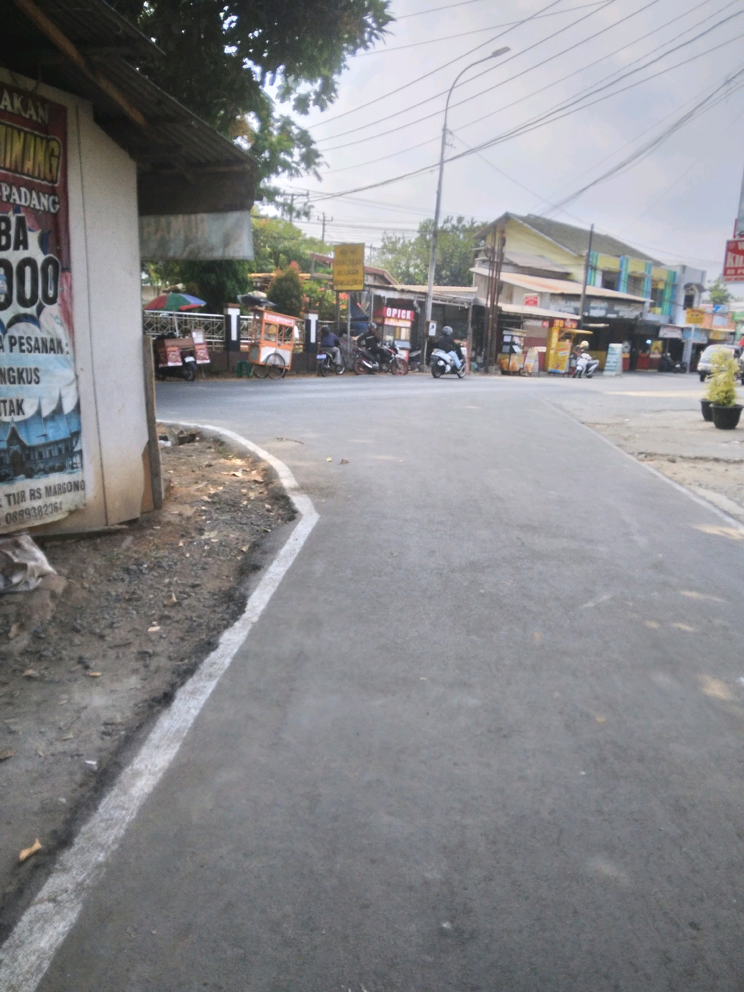The image shows a street scene in Desa Pamijen, Kecamatan Sokaraja, Indonesia. In the foreground, a gray asphalt road curves to the right, bordered by a white line on the left. Adjacent to the road is a patch of rough ground with dirt, gravel, and scattered leaves. A white-painted structure with a sloped, dark-colored roof stands on the left side of the frame. A portion of a large poster is visible on the side of this structure, featuring text and images.
Further down the street, several commercial stalls and buildings line the road. One stall has an orange and white cart with wheels. Several motorcycles are parked or being ridden. Two people are visible riding motorcycles, one on a white motorcycle and another on a darker motorcycle. A small tree or shrub is planted in a dark pot on the right side of the road. Overhead, a network of power lines is visible against a cloudy sky. The overall impression is a busy, possibly commercial area with a mix of vehicles and pedestrian activity.