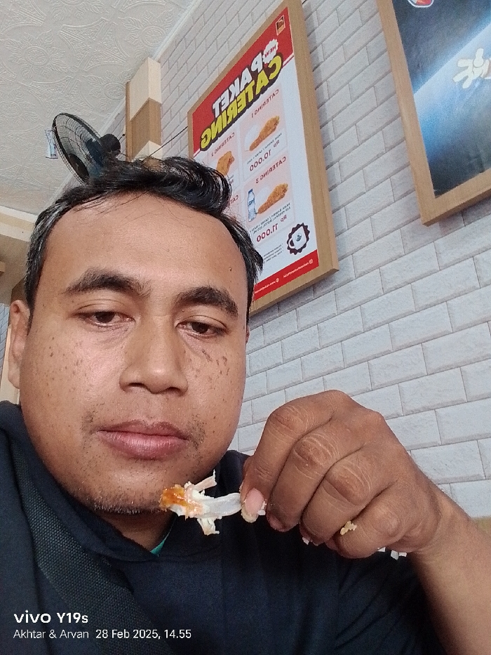 The image shows a man sitting in a restaurant in Patrol, Indonesia, located at latitude -6.318 and longitude 107.986. He is wearing a dark blue shirt and is holding a piece of fried chicken in his right hand. The restaurant has white brick walls with a red and white sign that reads "CATERING". The image appears to have been taken from a phone, as there is a reflection of a black object above the man's head. The image is dated February 28, 2025, at 14:55. The man is focused on the piece of chicken he is holding.