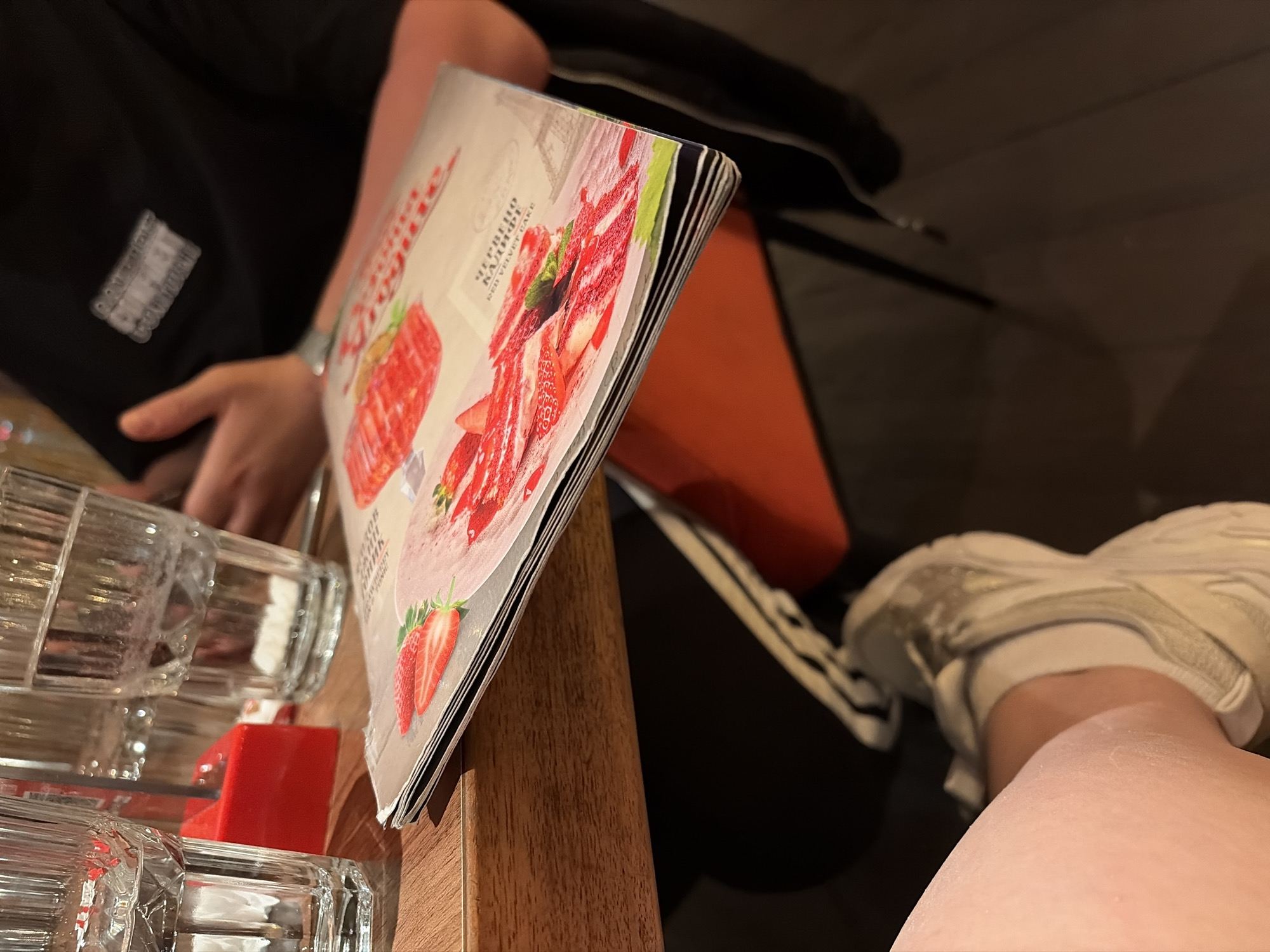 The image captures a close-up scene, likely indoors, with a partial view of a person seated. The person is wearing a black top with a white logo and white sneakers. A menu featuring images of red velvet cake and strawberries is visible and sitting on a light-brown wooden surface next to a rack of transparent glass bottles. A red divider is placed between the bottles. The location is potentially in Burgas, Bulgaria, indicated by the Cyrillic text on the menu, with the words "Червено кадифе" (Red Velvet).