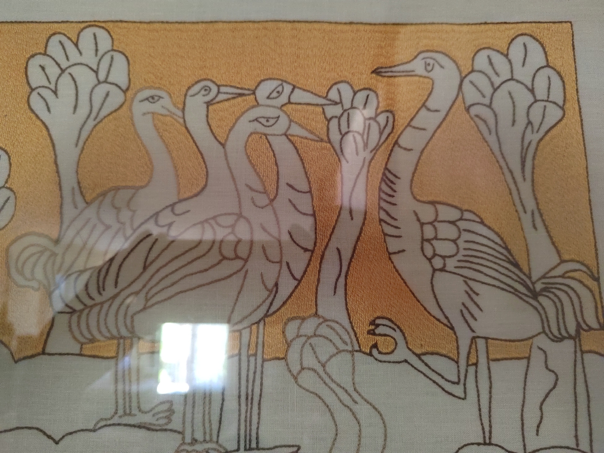 This is a close-up, eye-level, indoor shot of an embroidered textile featuring a stylized depiction of birds. The background is a textured gold fabric, while the birds and foreground elements are embroidered in outline stitch with a light beige thread on a white background. The birds appear to be cranes or herons, with long necks and legs, and distinctive fan-like shapes at the top of their bodies. Several birds are clustered together, with some necks intertwined. There are also abstract, leaf-like or floral elements on the left and right sides of the gold background. The embroidery style is simple and graphic, with minimal shading. A bright reflection of a window is visible on the lower left portion of the textile, obscuring some of the details in that area. The image is a photograph of the textile, likely framed or stretched. There is no indication of the city of Porto Recanati, Italy within the image or its context.