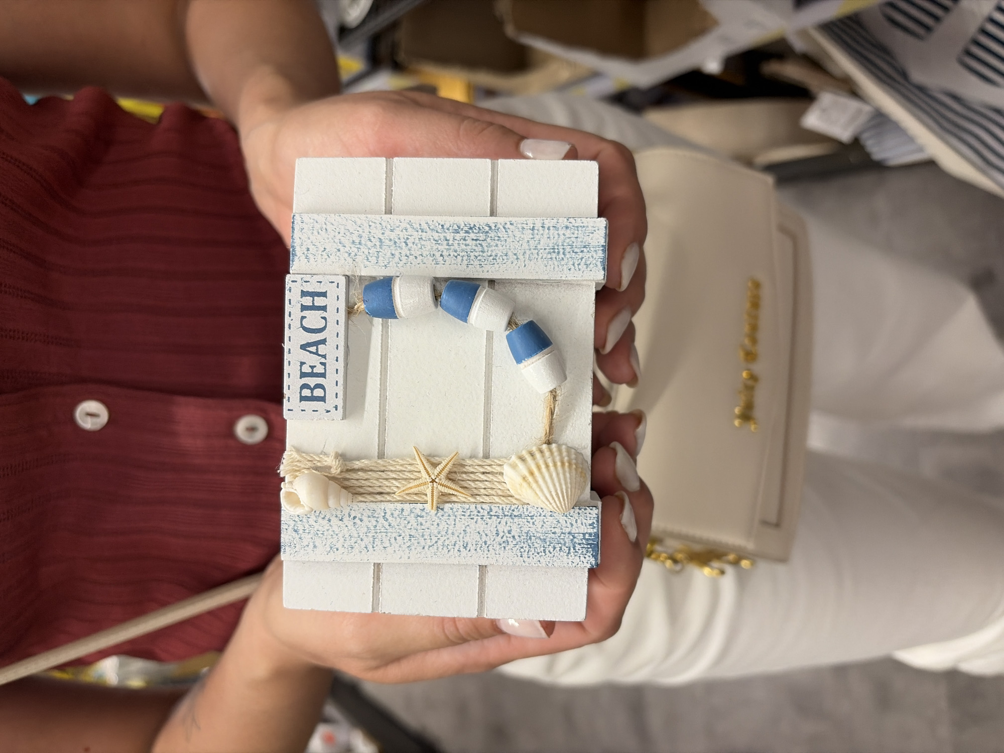 A person is holding a decorative sign that says "BEACH". The sign is made of white wood planks and decorated with shells, a starfish, and blue and white buoys. The person is wearing a maroon shirt and white pants. They are also carrying a small beige purse with gold lettering. The background is blurred, but it appears to be an indoor setting, possibly a store. There is no visible indication that this scene is located in Würzburg, Germany.