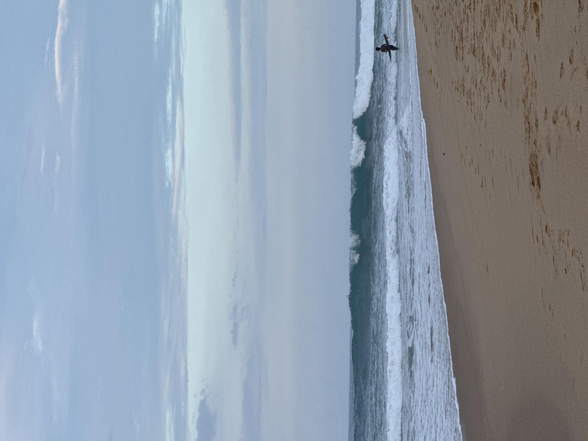 The image captures a serene beach scene in Arugam Bay, Sri Lanka. The foreground is dominated by a sandy beach, with subtle footprints suggesting recent activity. The ocean stretches out to the horizon, with moderate waves rolling towards the shore. A prominent wave is breaking in the mid-ground, its crest showing white foam. To the right of the breaking wave, a lone surfer stands in the shallow water, holding a surfboard. The surfer is facing the ocean, appearing to be waiting for a suitable wave. The sky above is a soft, muted blue, with scattered clouds adding a gentle texture to the atmosphere. The overall impression is one of calm and tranquility, characteristic of a peaceful day at the beach.