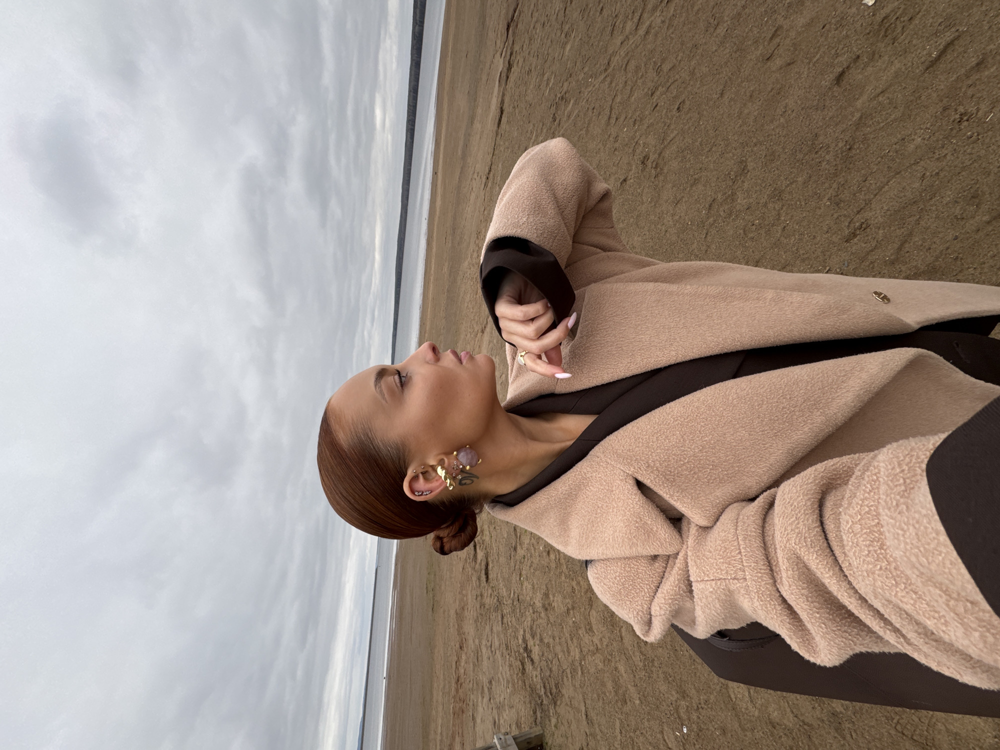 A woman is standing on a sandy beach. Behind her is a body of water with a strip of land visible on the opposite shore. The sky is overcast with a gray cloud cover. The woman is wearing a tan coat over a dark-colored garment. Her hair is pulled back in a bun, and she is wearing ornate earrings. Her right hand is raised. The location is Chaikovsky, Russia.