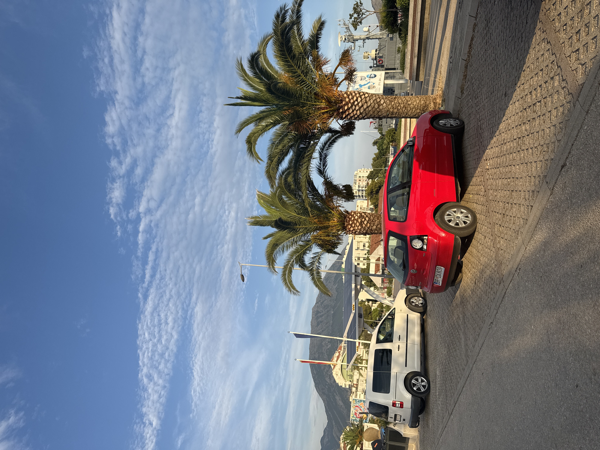 The image shows a street scene in Bar, Montenegro, with two parked cars in the foreground: a white van and a red hatchback. The van is on the left, facing right, and the red car is on the right, facing left. Both vehicles are parked on a textured paved surface, possibly a pedestrian walkway or parking area. Behind the cars, several palm trees are visible, with their fronds creating a partial canopy. In the background, there are buildings, mountains, a solar panel installation, and a ship docked at a harbor. The sky is blue with scattered white clouds, suggesting it is daytime. The overall impression is a sunny, coastal environment.