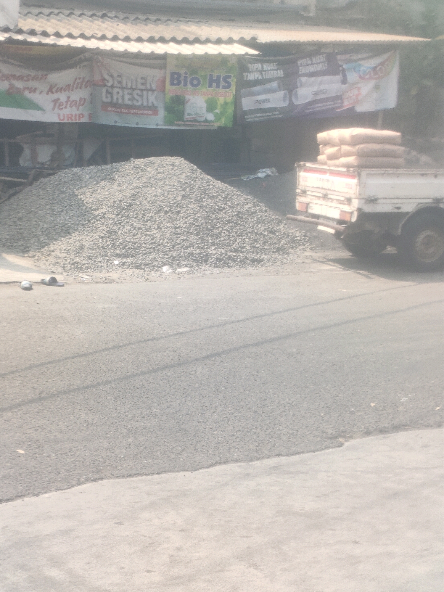 The image depicts a street scene in Surabaya, Indonesia, characterized by a construction supply area. In the background, a covered awning displays several banners advertising various building materials. One banner prominently features "SEMEN GRESIK," a well-known Indonesian cement brand, alongside other advertisements for pipes and construction-related products.
In the foreground, a large pile of gravel or crushed stone occupies a significant portion of the frame, indicating active construction or material storage. To the right of the gravel pile, a small white truck is parked, loaded with several sacks of material, likely cement. The truck has its taillights visible and appears to be a flatbed type.
The ground is a paved road, with a clear transition to what appears to be a lighter-colored concrete surface or sidewalk on the right side of the image. The overall lighting suggests it is daytime, possibly late morning or afternoon, with a hazy or dusty atmosphere that softens the details. Two pairs of slippers are visible on the ground near the edge of the gravel pile. The scene appears to be an outdoor commercial area, likely a building materials vendor.