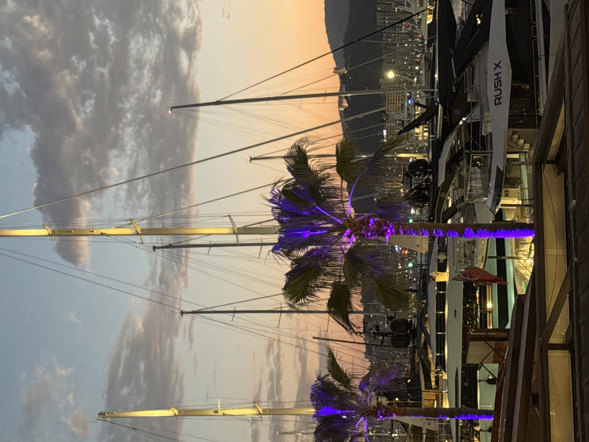 The image depicts a marina in Palma, Spain at dusk. Several yachts and sailboats are moored in the harbor, with their masts visible against the twilight sky. Two palm trees, illuminated with purple lights, stand in the foreground. In the background, a cityscape with buildings and lights is visible, nestled against a hillside. A red flag with a white cross is seen flying from one of the yachts. The sky is a gradient of pale blue and orange, with scattered clouds. The overall scene conveys a sense of luxury and tranquility.