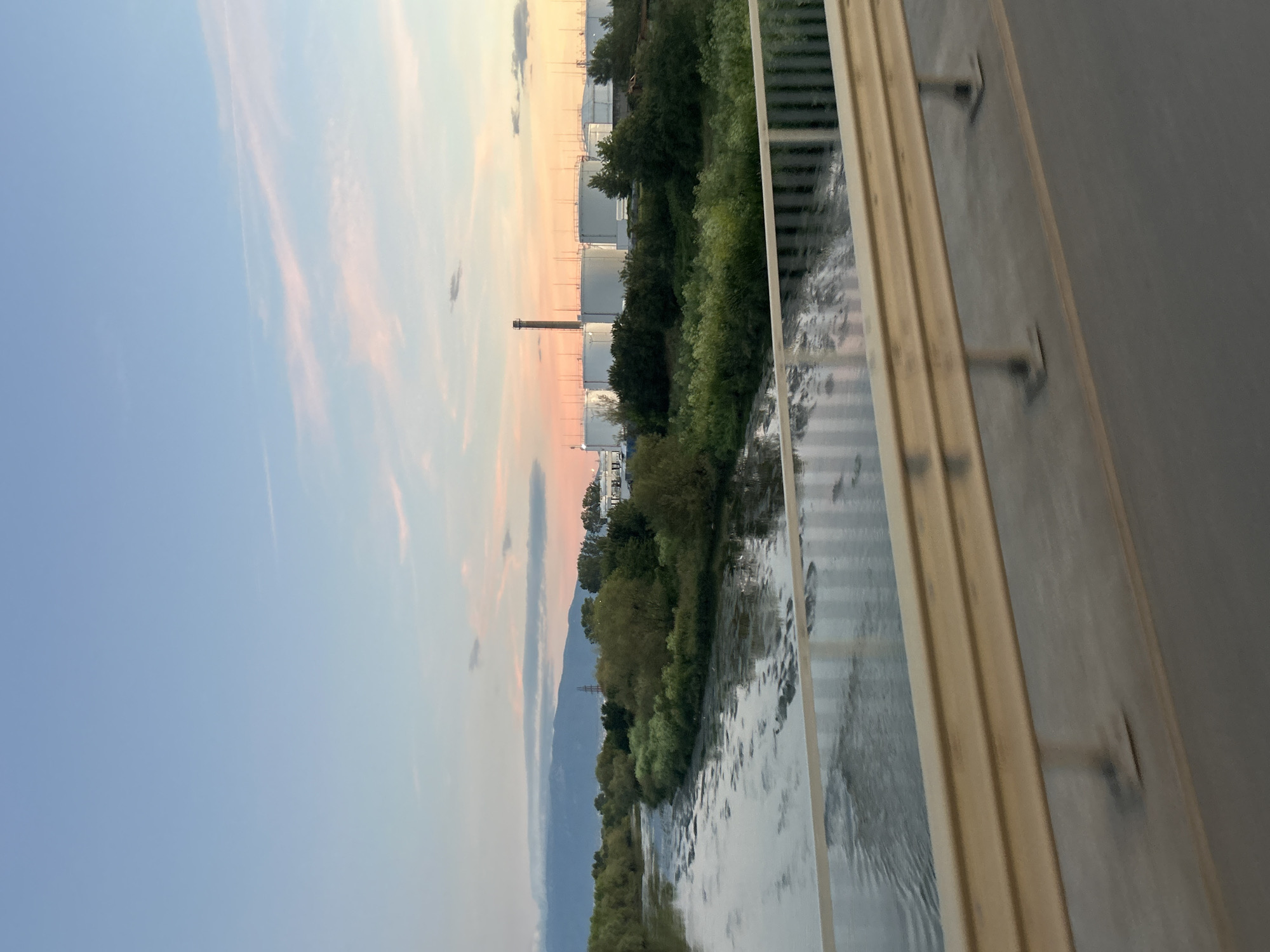 An outdoor, eye-level, horizontal shot captures a wide river flowing from the left to the right of the frame. The sky above is a soft gradient of blue and pink, indicating it might be dawn or dusk. In the background, a row of large, white cylindrical tanks, likely part of an industrial facility, is visible behind a lush green tree line. A tall, slender smokestack rises from the industrial area. To the left, distant mountains are shrouded in a light haze. In the foreground, a yellow guardrail extends across the bottom of the image, separating the viewer from the river below. The water appears murky with reflections of the sky and vegetation.