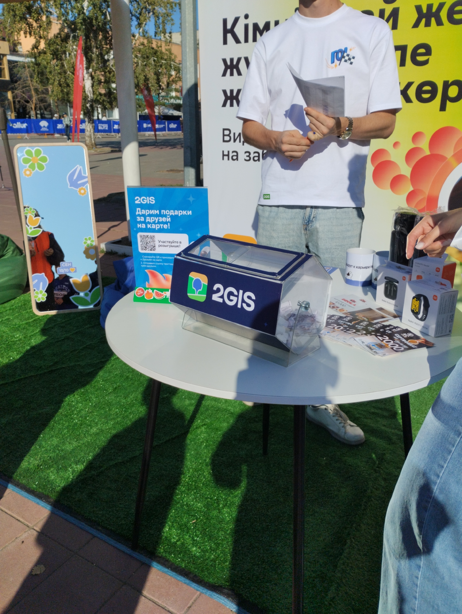 The image captures an outdoor promotional event, possibly a lottery or giveaway, set up on artificial turf in a city. A white table is centrally positioned, displaying promotional materials and a clear raffle drum filled with slips of paper. To the left, a tall, rectangular display board features colorful floral graphics and a reflection of a person in orange and grey clothing. A blue sign with the 2GIS logo and promotional text in Russian, "Дарим подарки за друзей на карте!" (We give gifts for friends on the map!), along with a QR code, is placed near the raffle drum.
In the background, a person in a white t-shirt with a checkered logo and the text "10%OFF" is holding a document and wearing a wristwatch. Behind them, a large yellow banner with large black Russian lettering, including "Кім" (Kim) and "же" (zhe), is visible. The environment suggests an urban setting with trees and buildings in the distance. The event appears to be taking place on a sunny day, as indicated by the shadows. The specific location in Kostanay, Kazakhstan, is not definitively identifiable from the visual cues alone, but the presence of Russian language signage might indicate a connection to a Russian-speaking region or a company operating in such areas.